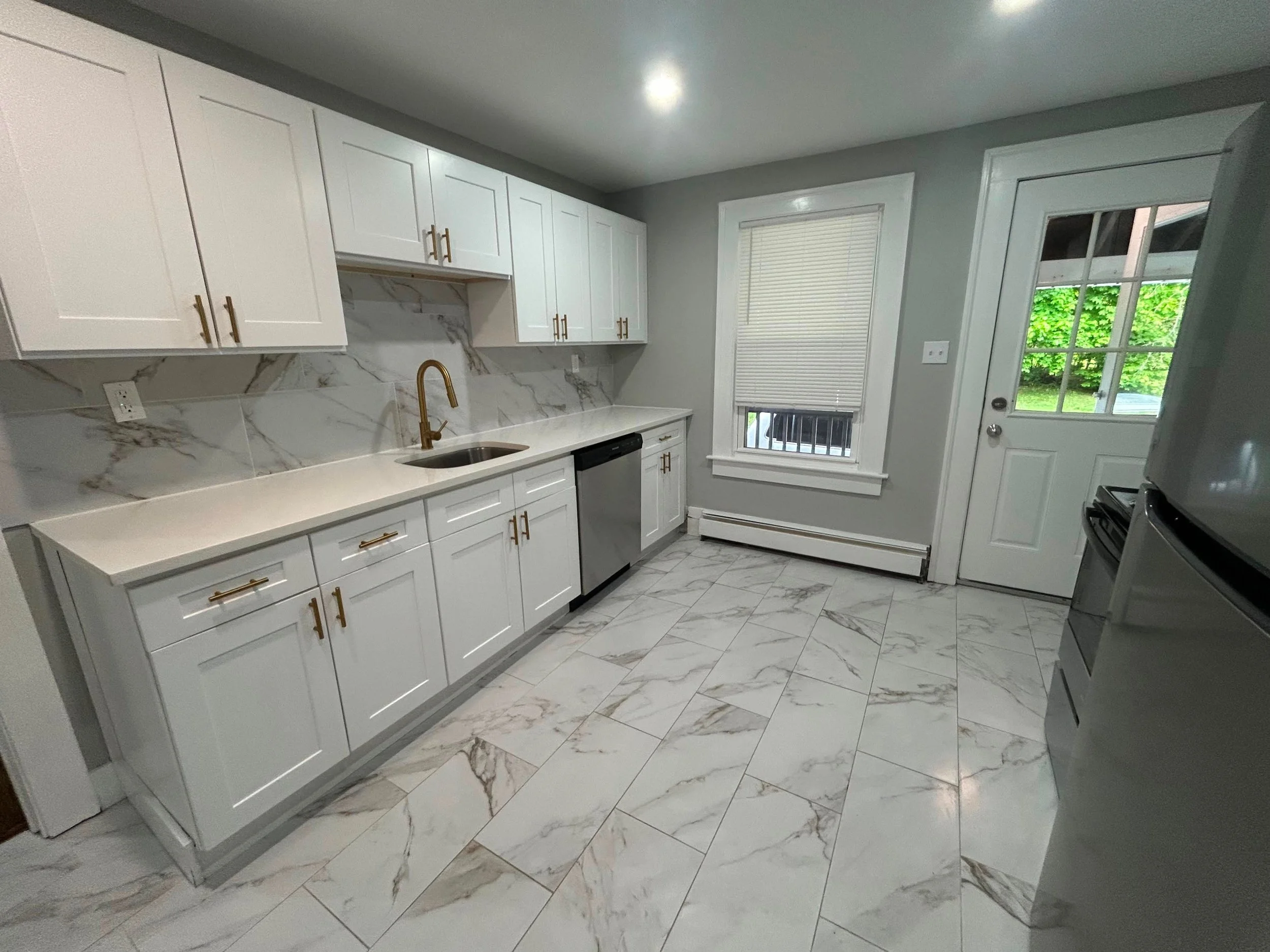 A modern kitchen with white cabinets, marble backsplash, marble floor tiles, a window with closed blinds, a door with a window, and a gold faucet.