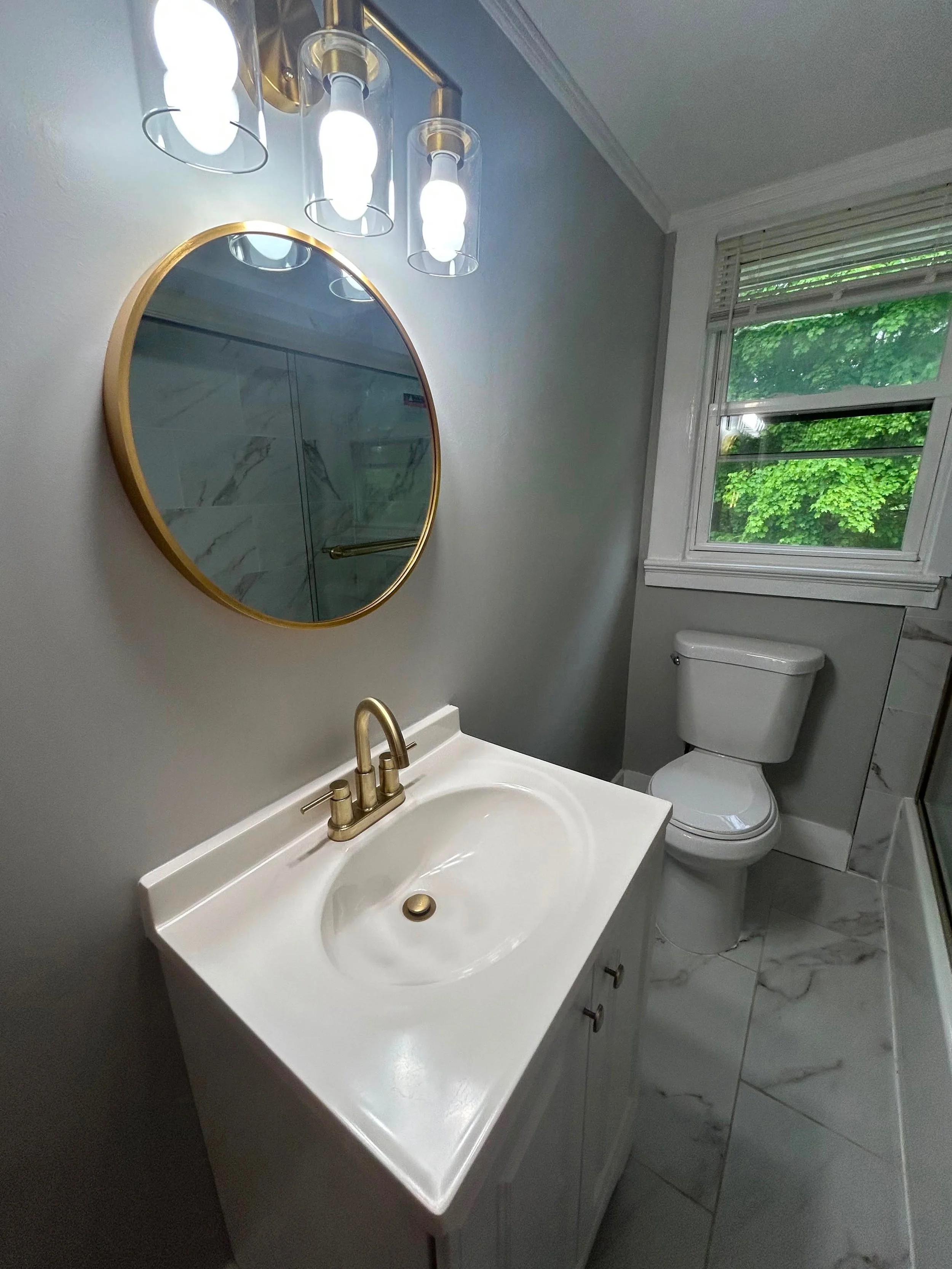 A small bathroom with a white sink, gold faucet, oval mirror with gold frame, and a window showing green trees outside.