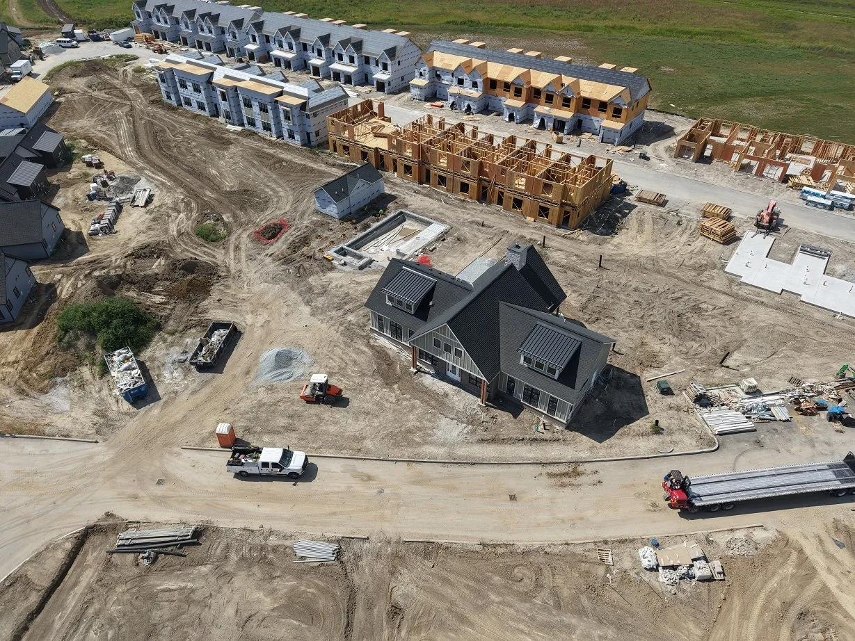 Aerial view of a residential construction site with multiple houses in different stages of development, including partially built wooden frames, finished houses, construction vehicles, and building materials.