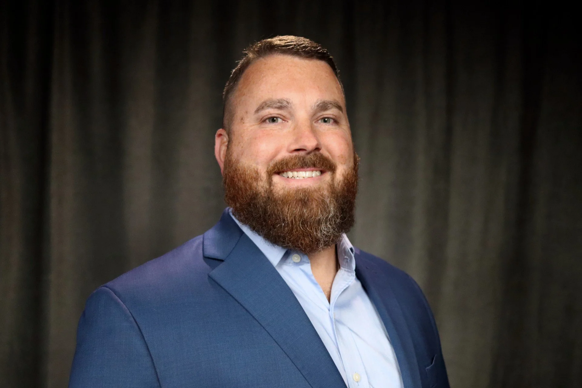 A man with a beard and short hair wearing a blue suit and light blue shirt, smiling at the camera, with a dark curtain backdrop.
