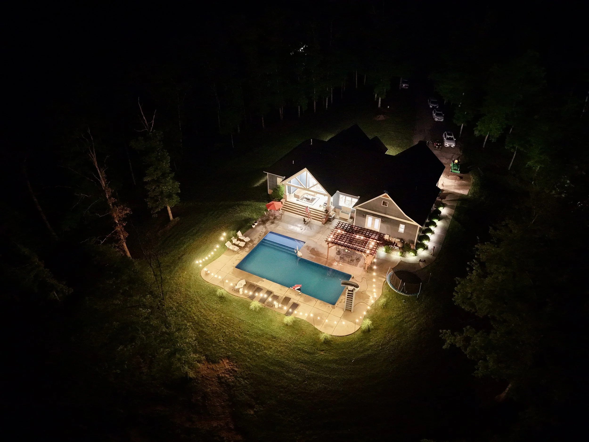 Aerial night view of a house with a lit backyard pool, outdoor seating, and string lights, surrounded by trees and parked cars.