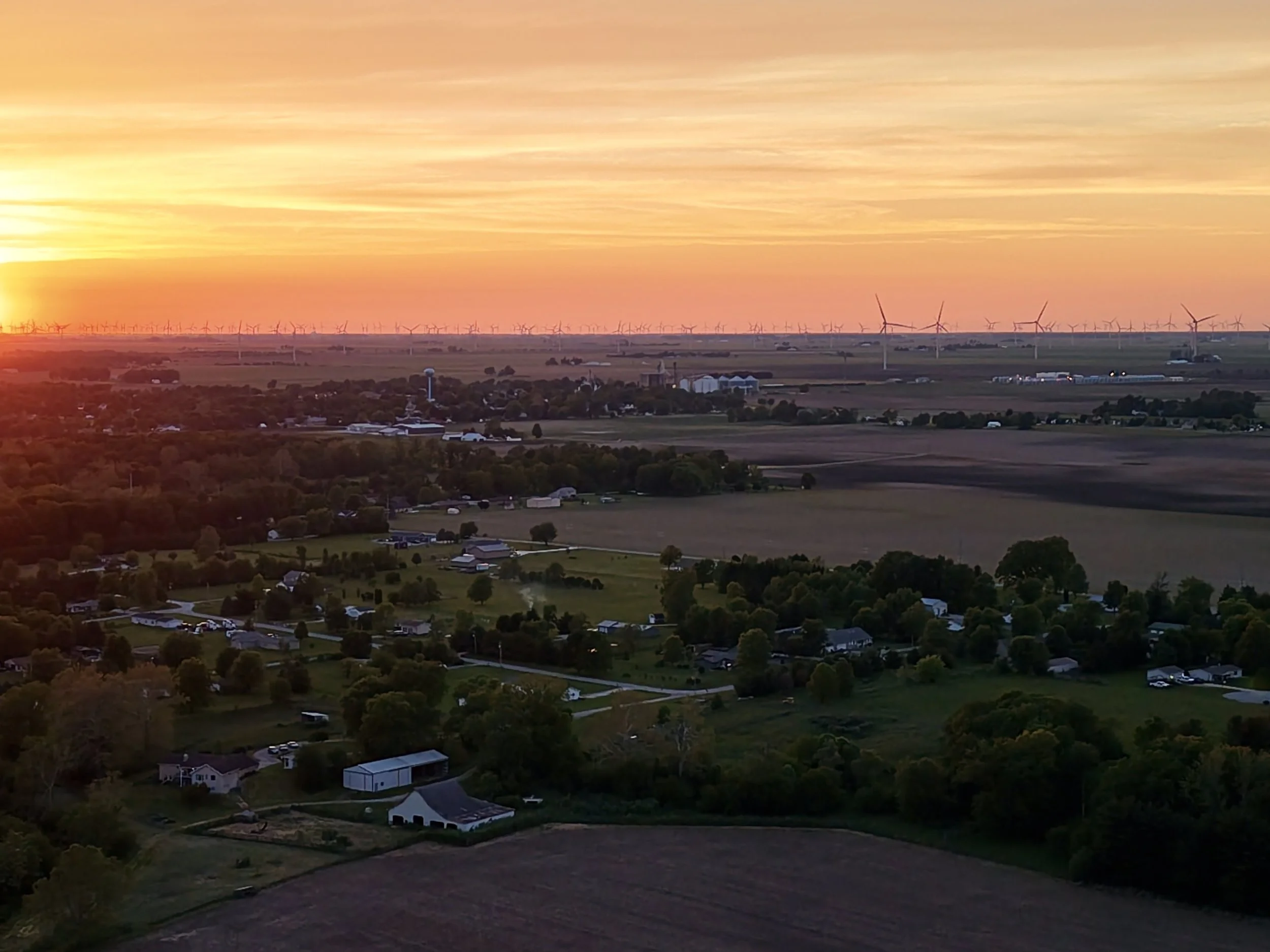 A rural landscape during sunset with numerous wind turbines on the horizon, scattered houses, trees, and open fields.