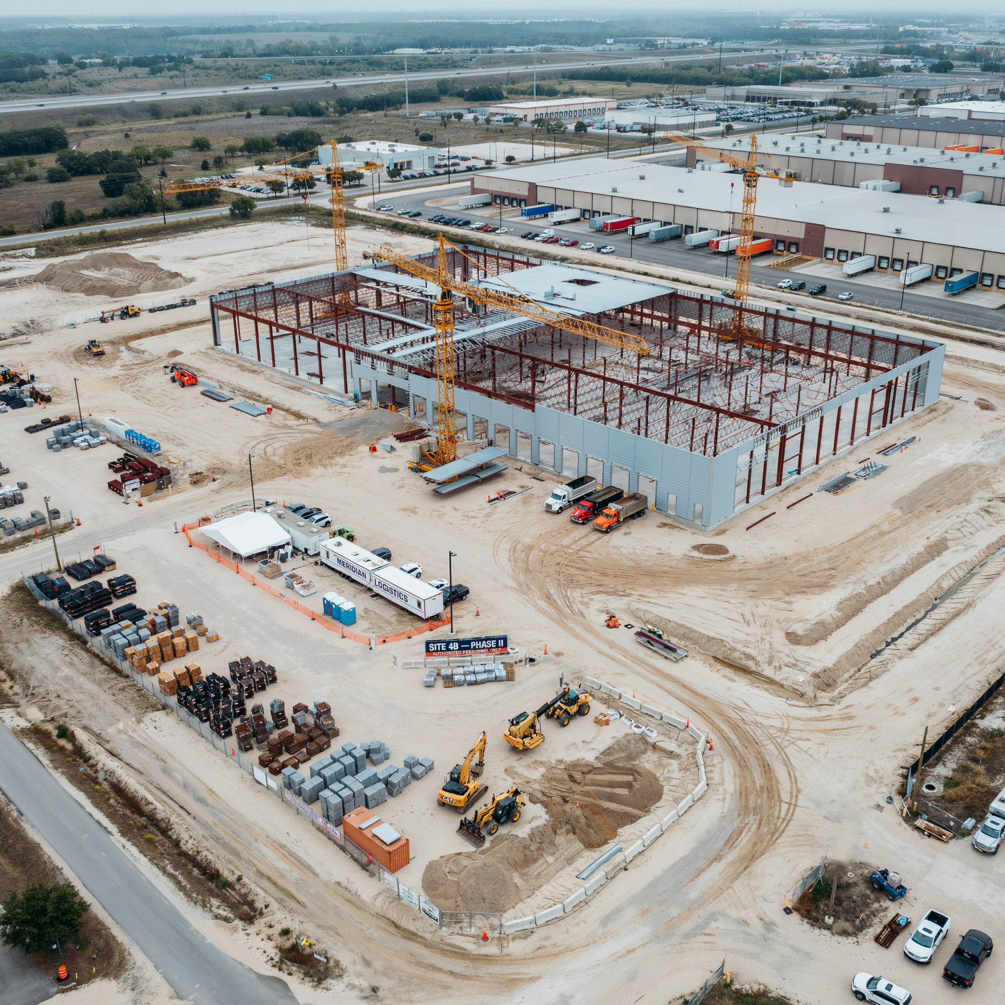 Aerial view of a commercial construction site with a partially built structure, yellow cranes, construction vehicles, and surrounding parking lots with trucks and cars.