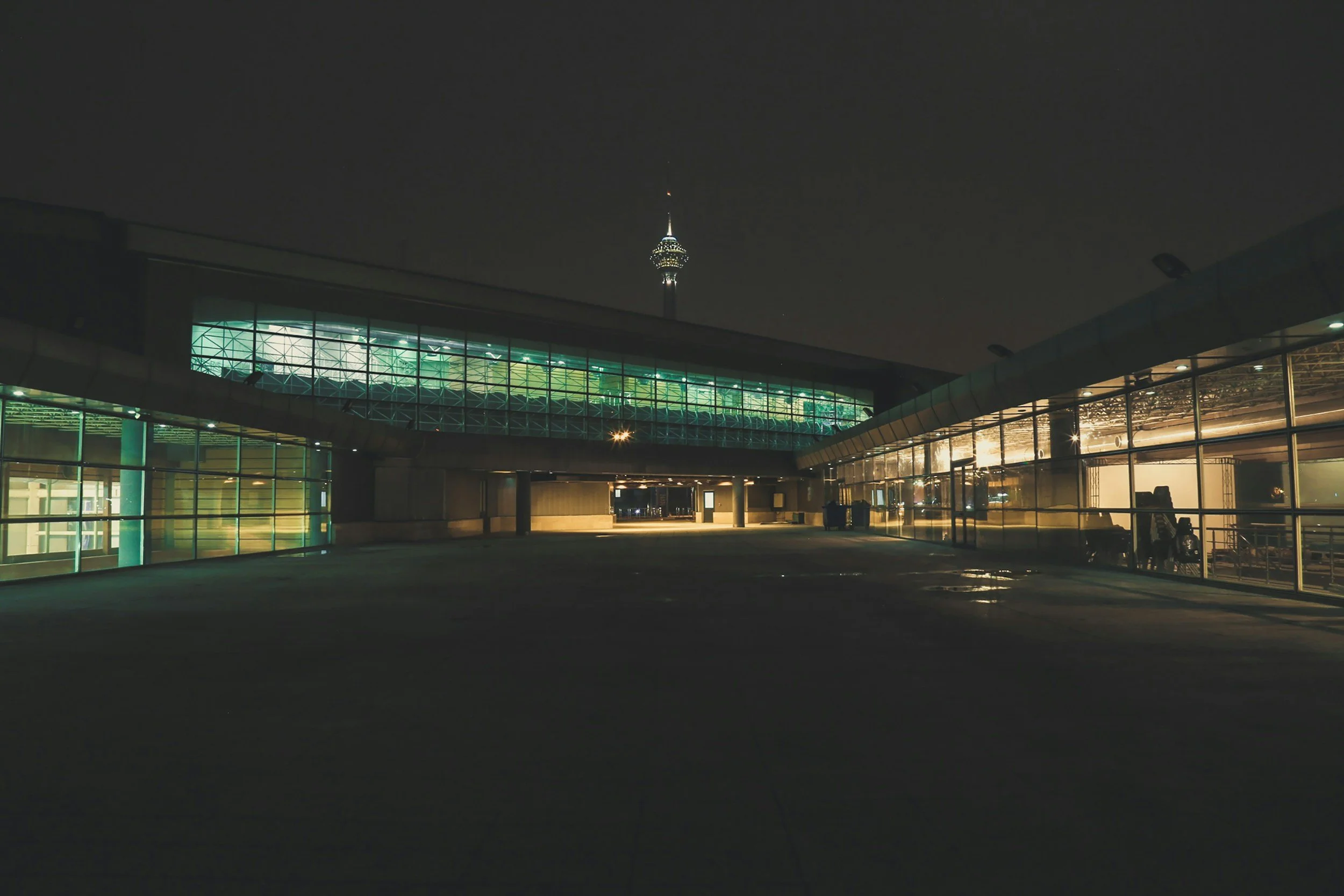 Nighttime view of an airport terminal with glass walls, illuminated from within, and a tall tower visible in the background. The sky is dark, and the area appears quiet with no visible people.