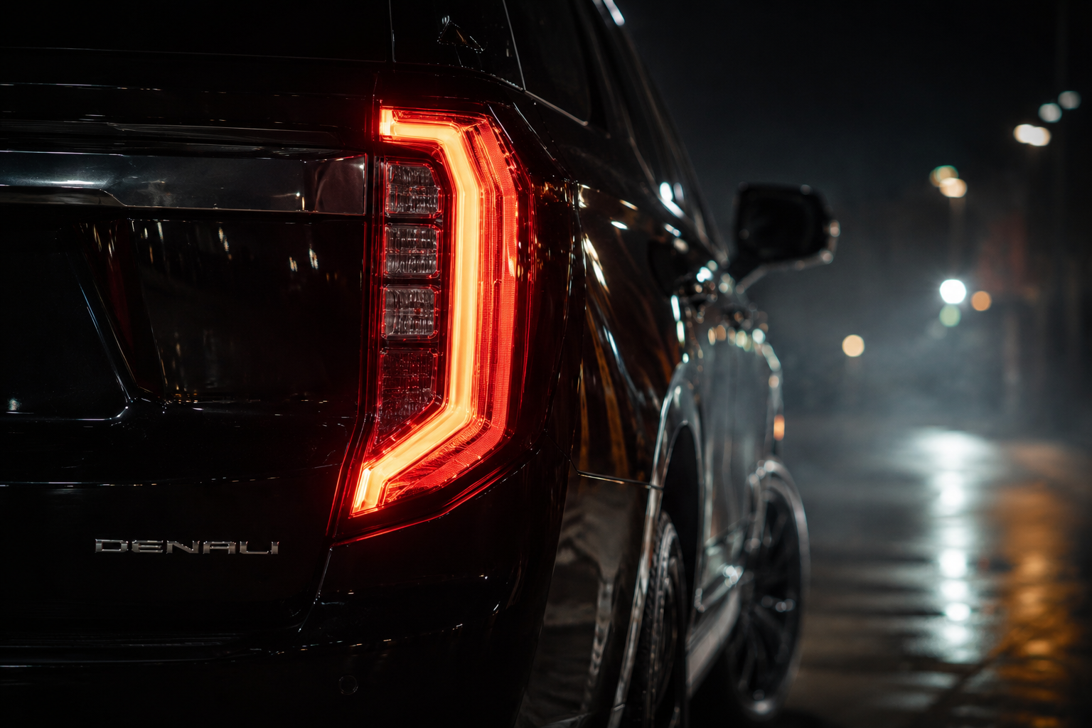 Close-up of the rear left side of a black GMC Yukon Denali at night, showing the illuminated taillight and a wet road surface.