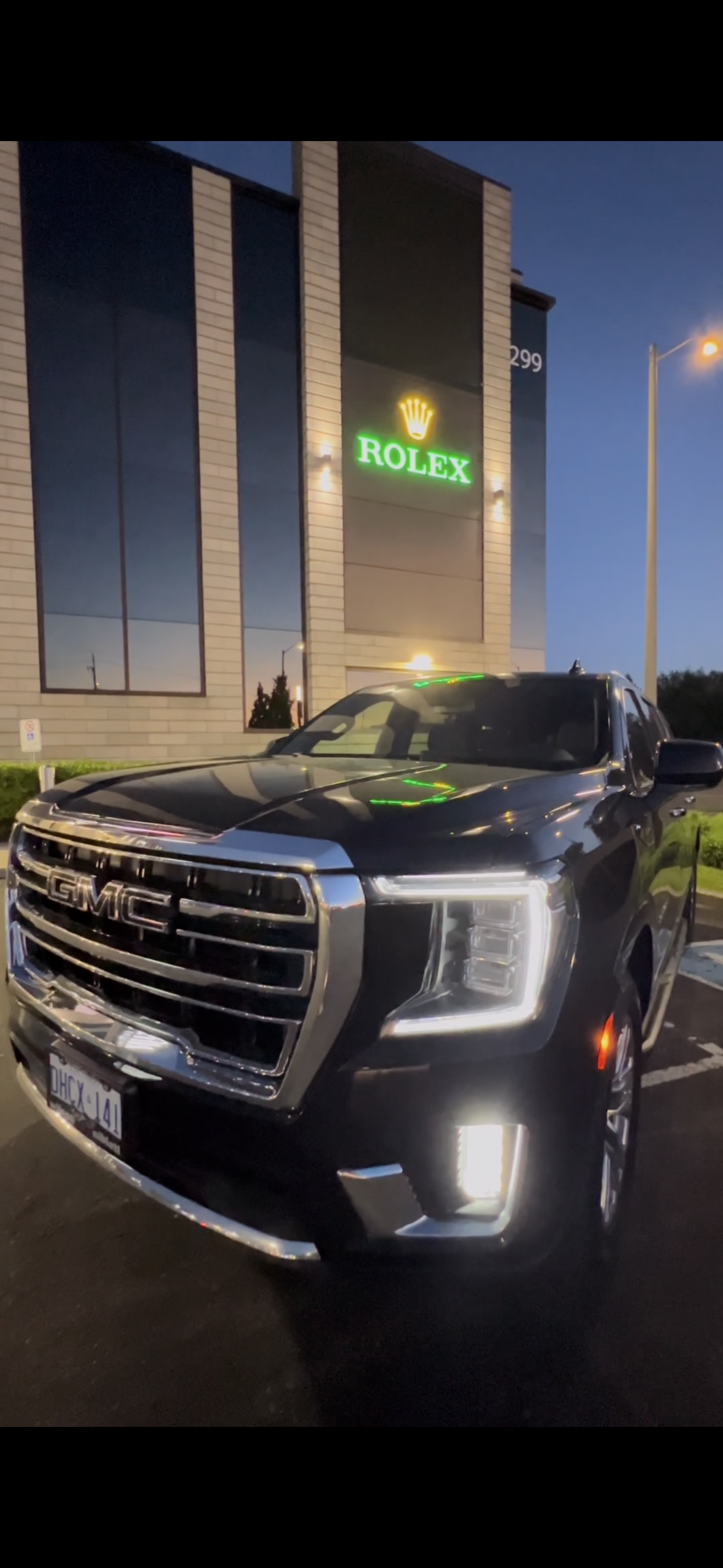 Black GMC SUV parked in front of a building with a neon Rolex sign at dusk.