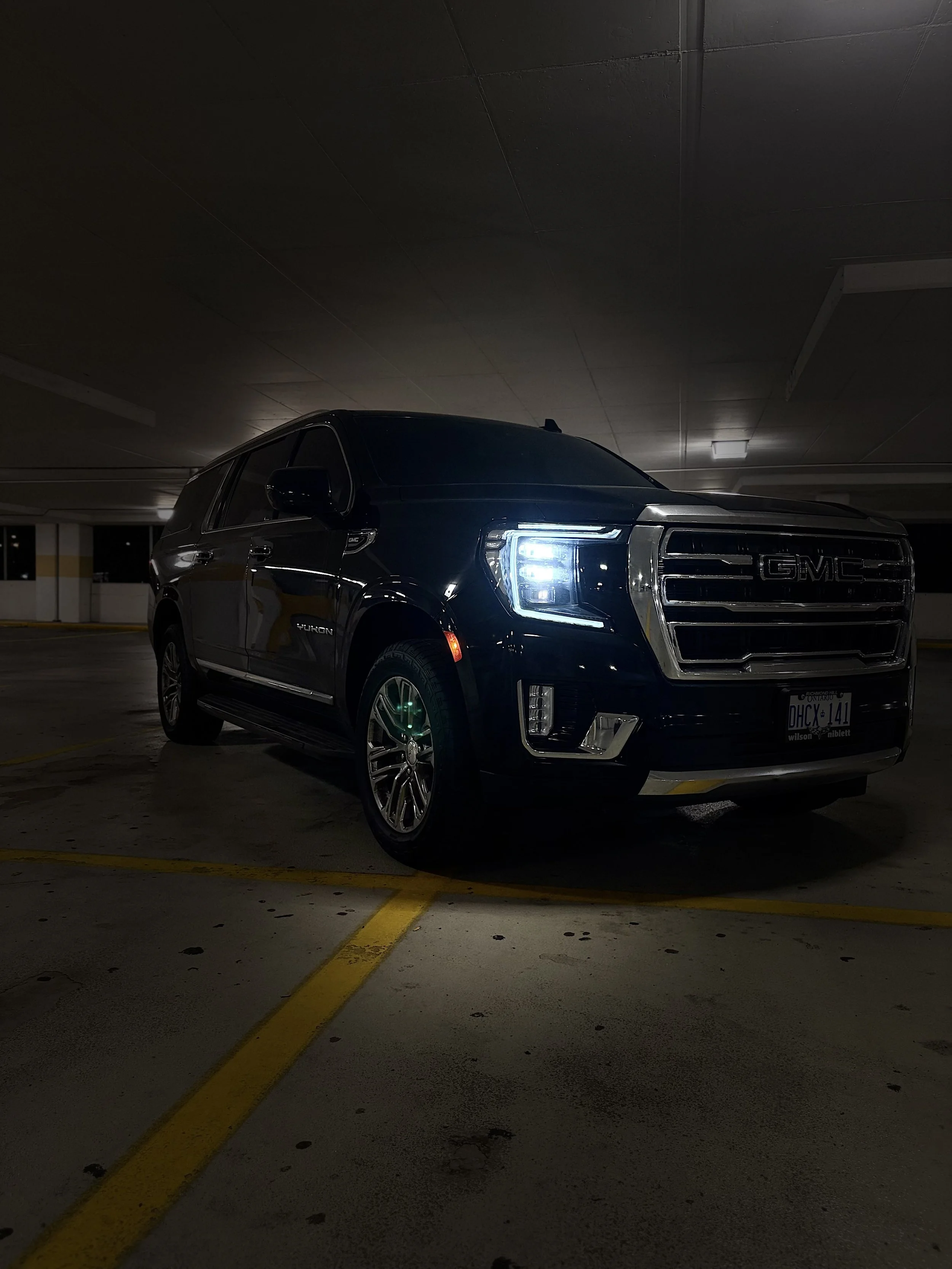 A black GMC Yukon parked in an underground parking lot at night, with its headlights on and yellow parking lines visible on the concrete floor.