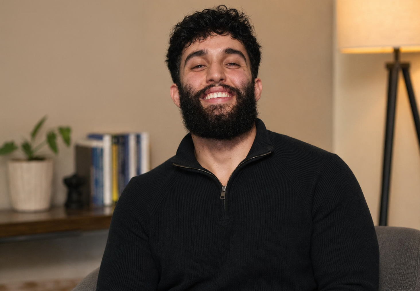 A smiling man with curly dark hair and a full beard, wearing a black zip-up sweater, sitting in a room with a beige wall, a potted plant, books, and a lamp in the background.