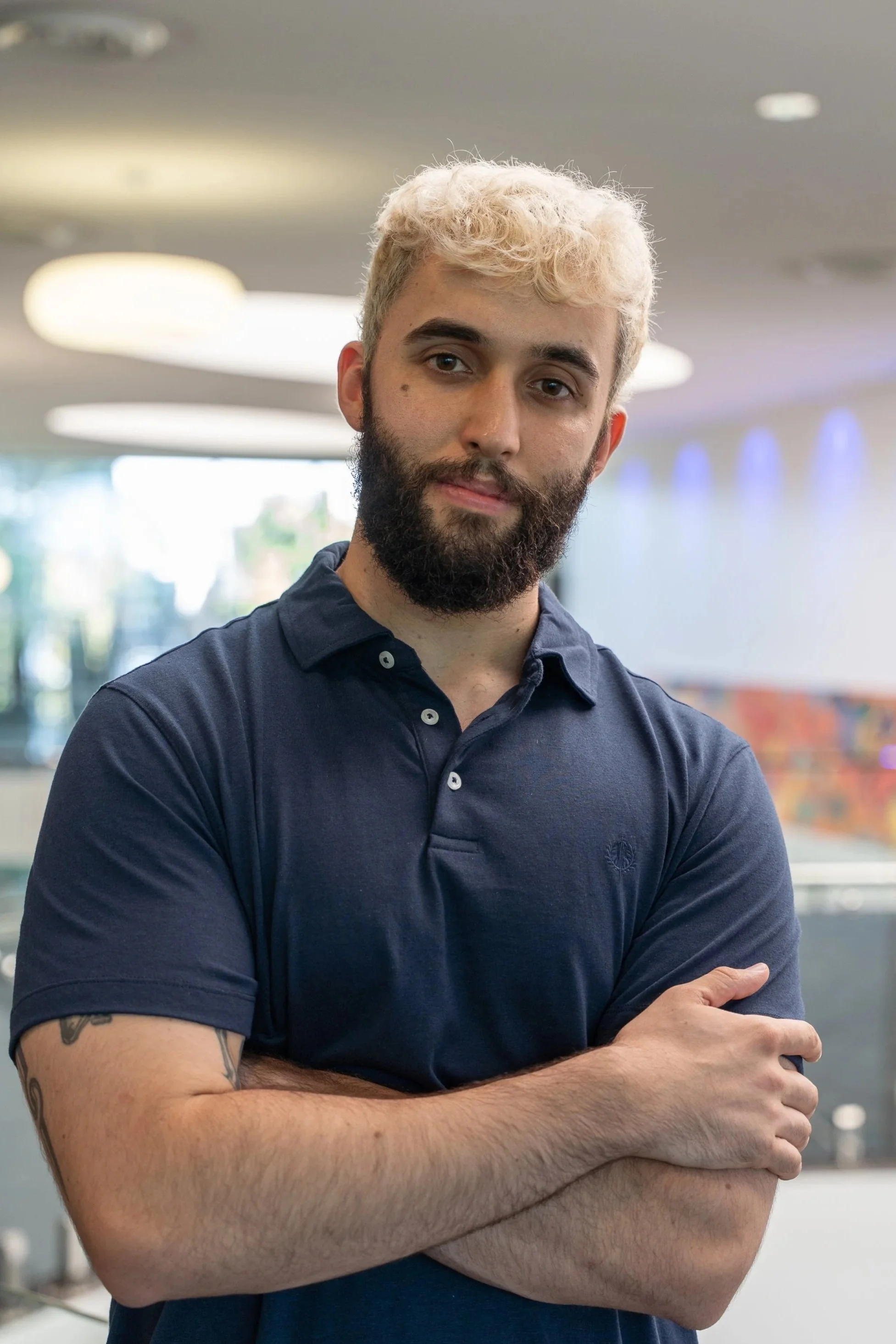 A young man with curly blond hair and a beard, wearing a navy polo shirt, standing indoors with his arms crossed, in a modern setting with blurred lights and windows in the background.