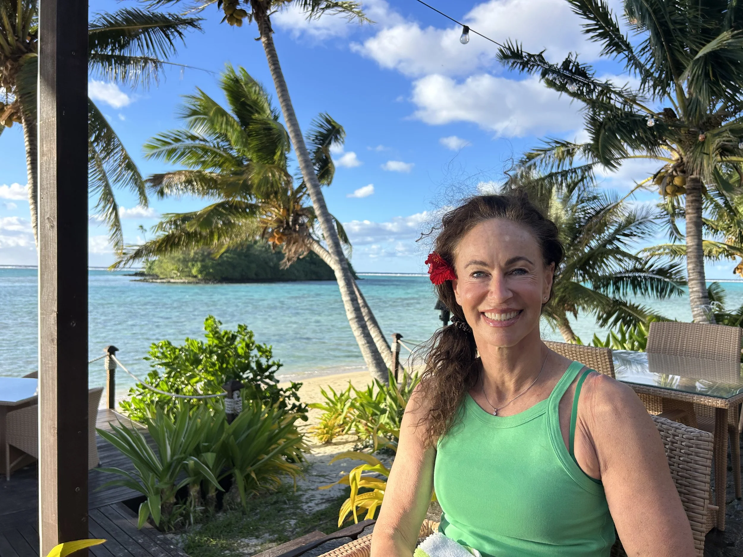 Anandi with a red flower in her hair wearing a green tank top taking a selfie on a beachside patio with palm trees, blue sky, ocean, and island in the background, under string lights.