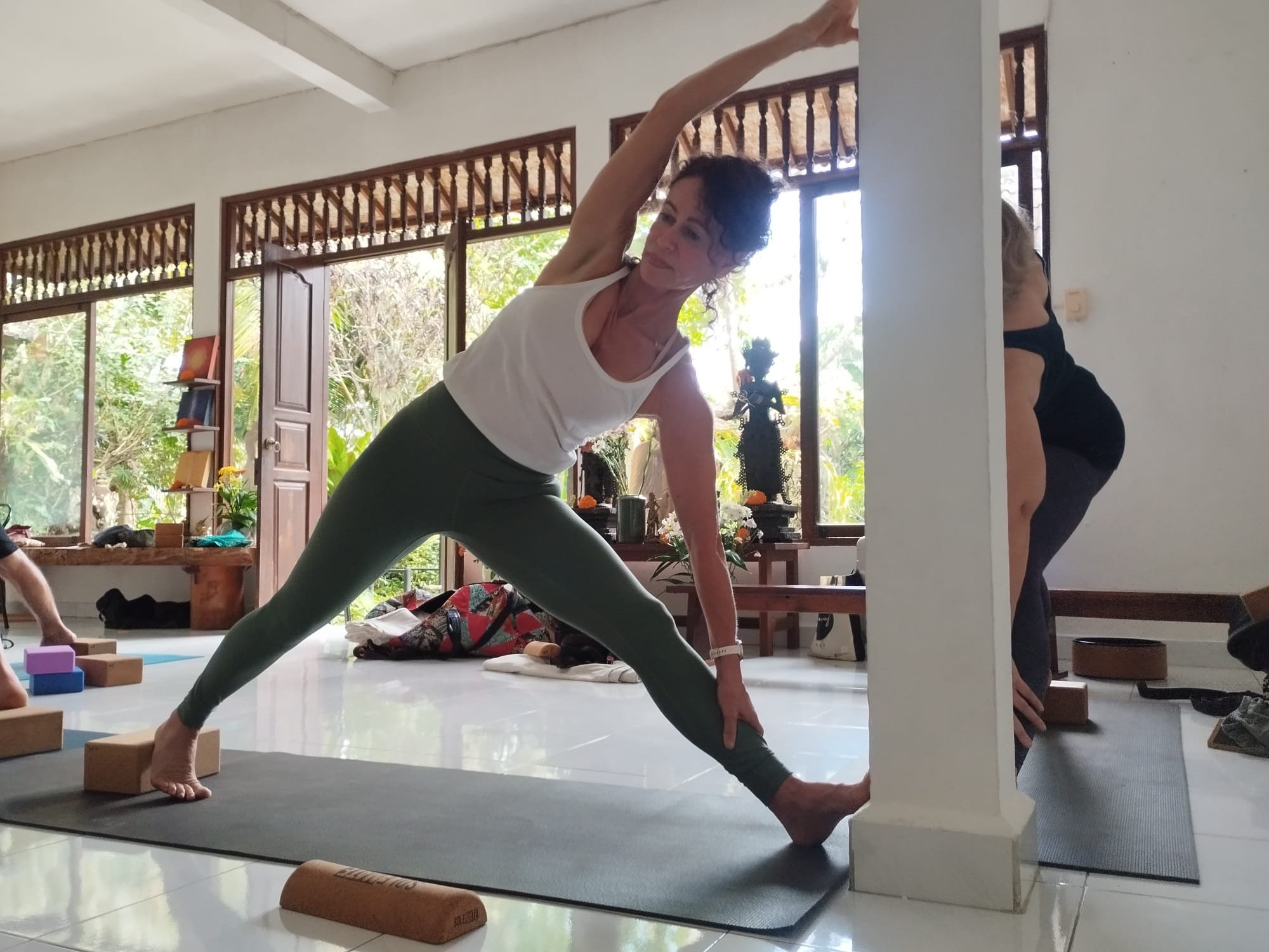 Anandi practicing yoga in a bright room with large windows, plants, and wooden decor, performing a side angle stretch.