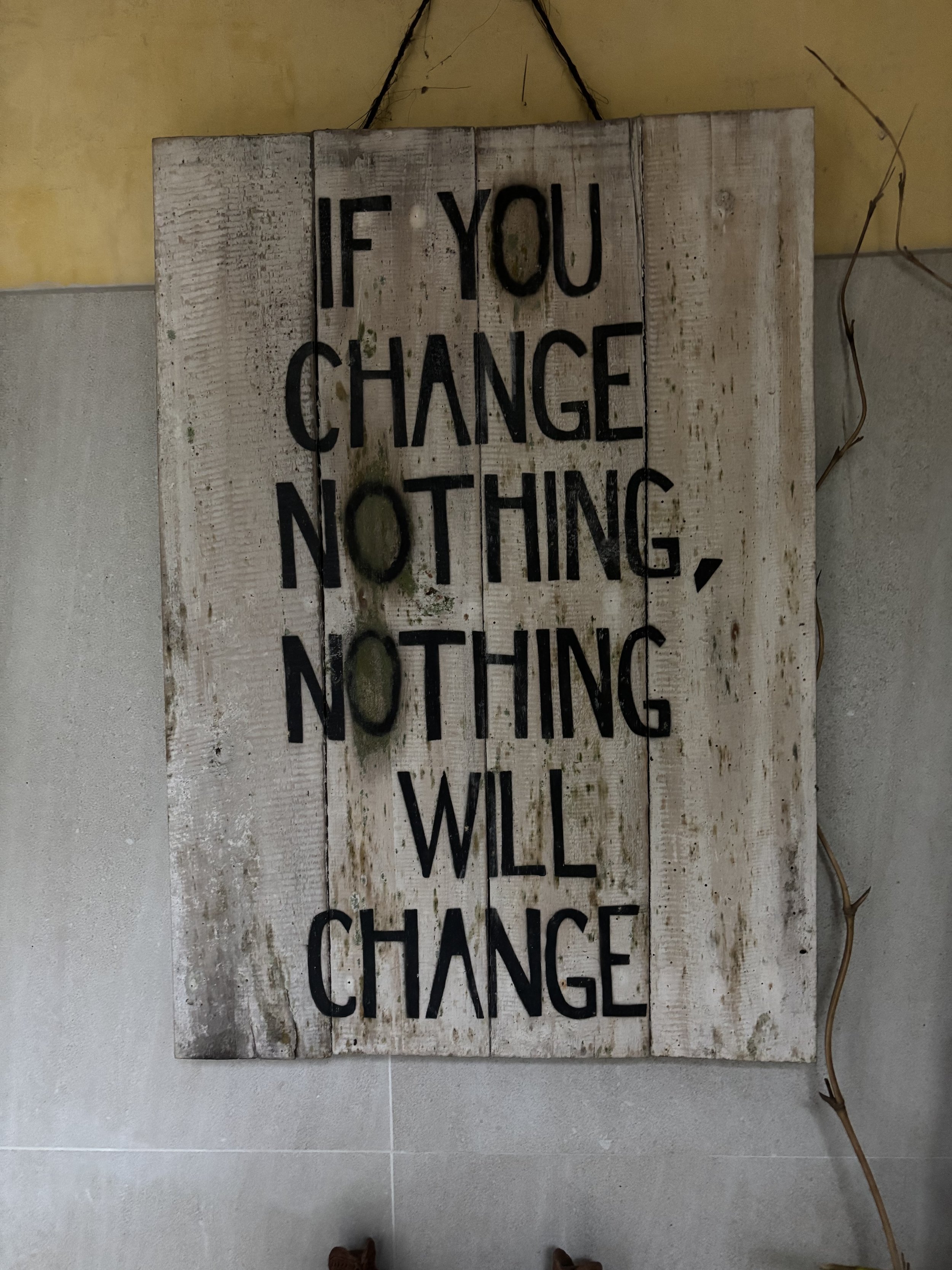 A rustic wooden sign with black lettering that reads "If you change nothing, nothing will change". The sign is hanging on a beige wall with electrical wires visible on the right side.