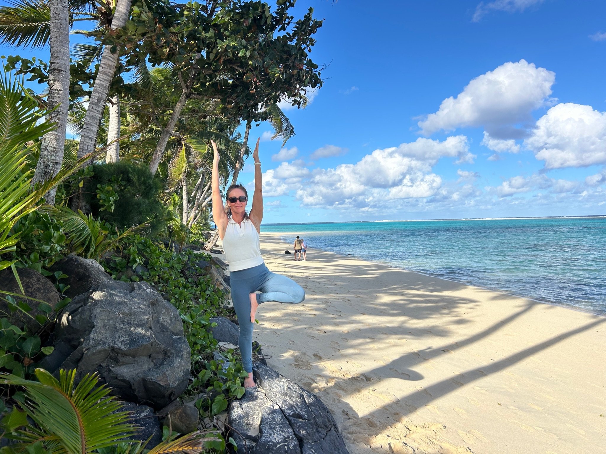 A woman in casual clothes and sunglasses practicing yoga in a tree pose on a tropical beach with clear blue water, sandy shore, lush greenery, and a partly cloudy sky.