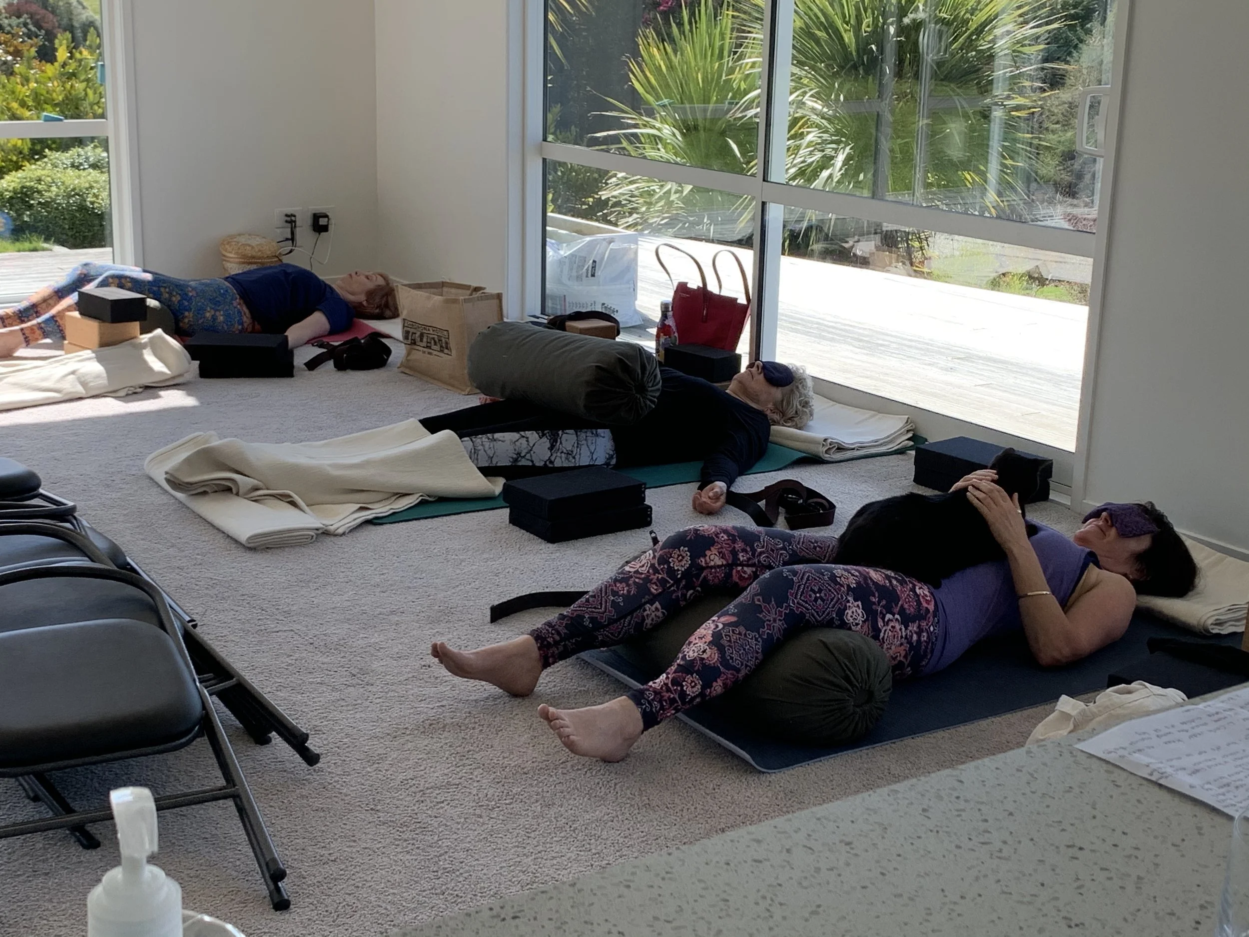 Several people are lying on yoga mats and blankets on the floor of a room with large windows showing a garden with green plants. It appears to be a yoga or relaxation class.