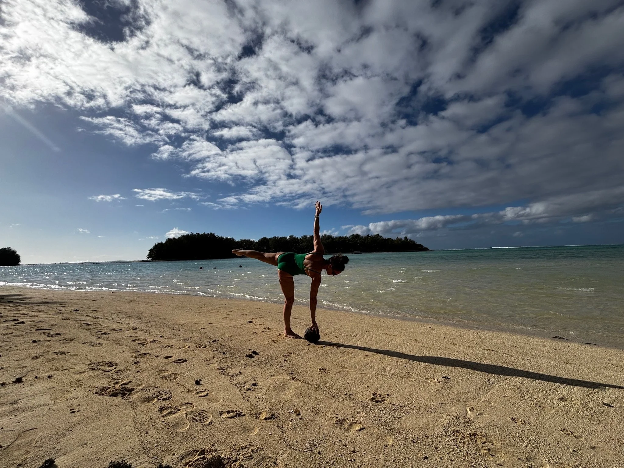 Person practicing handstand yoga pose on a sandy beach near the ocean with cloudy sky and trees in the background.
