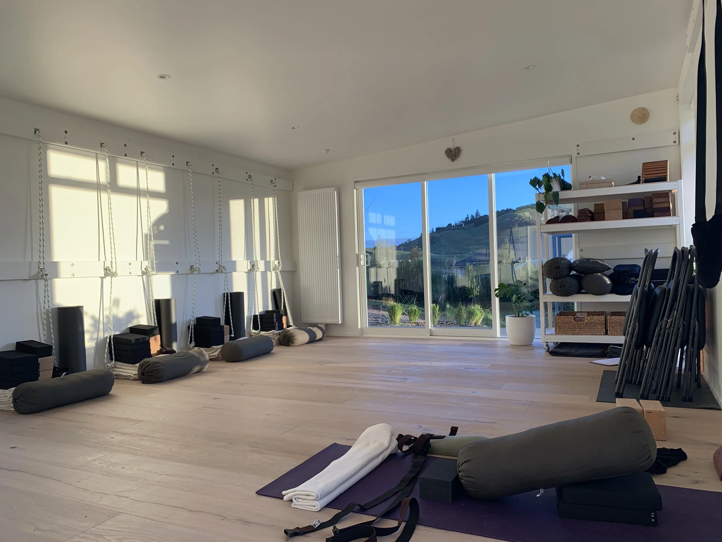 A spacious yoga or exercise room with large windows revealing a view of green hills and blue sky, white walls with ropes and padded swings, and neatly arranged mats and props for practice.