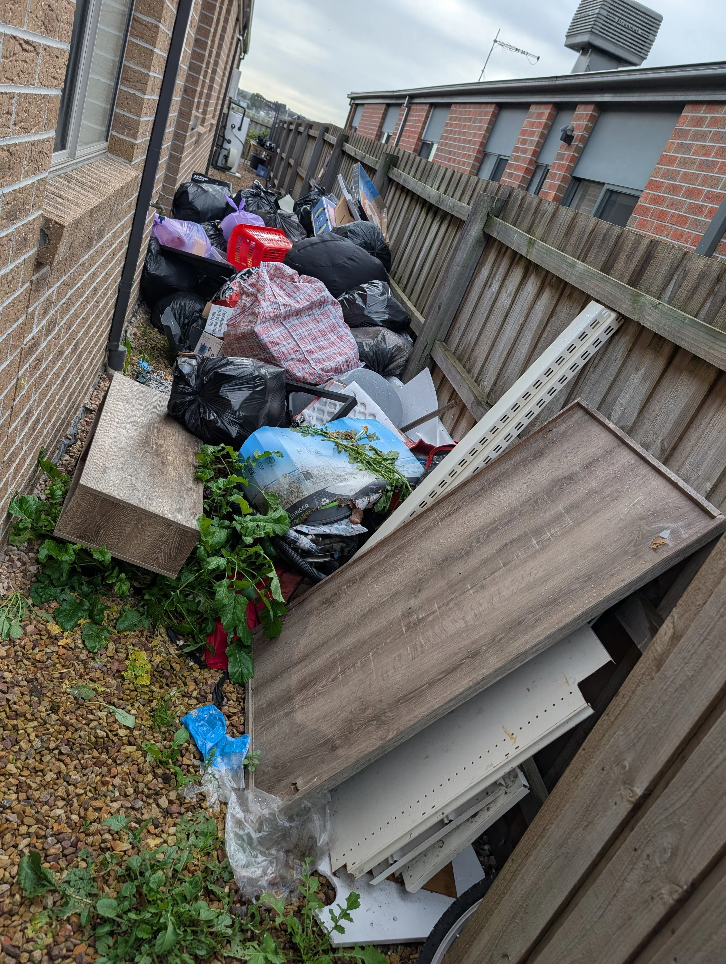 Accumulation of trash including black garbage bags, cardboard boxes, plastic containers, and miscellaneous debris along a wooden fence outside a house with a brick exterior.