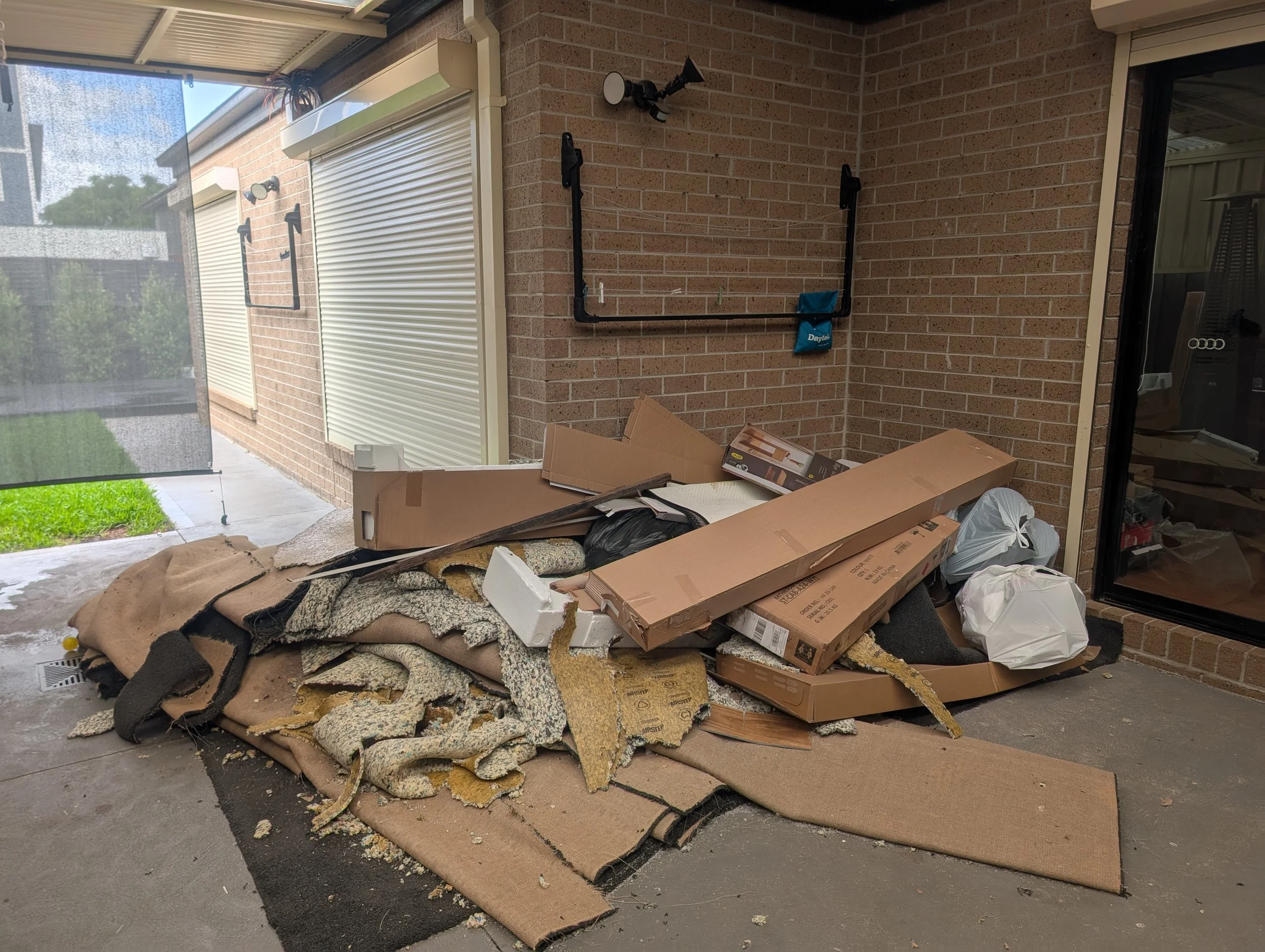 Piles of insulation foam and cardboard boxes on a concrete floor in a covered outdoor area next to a house with brick walls and closed white shutters.