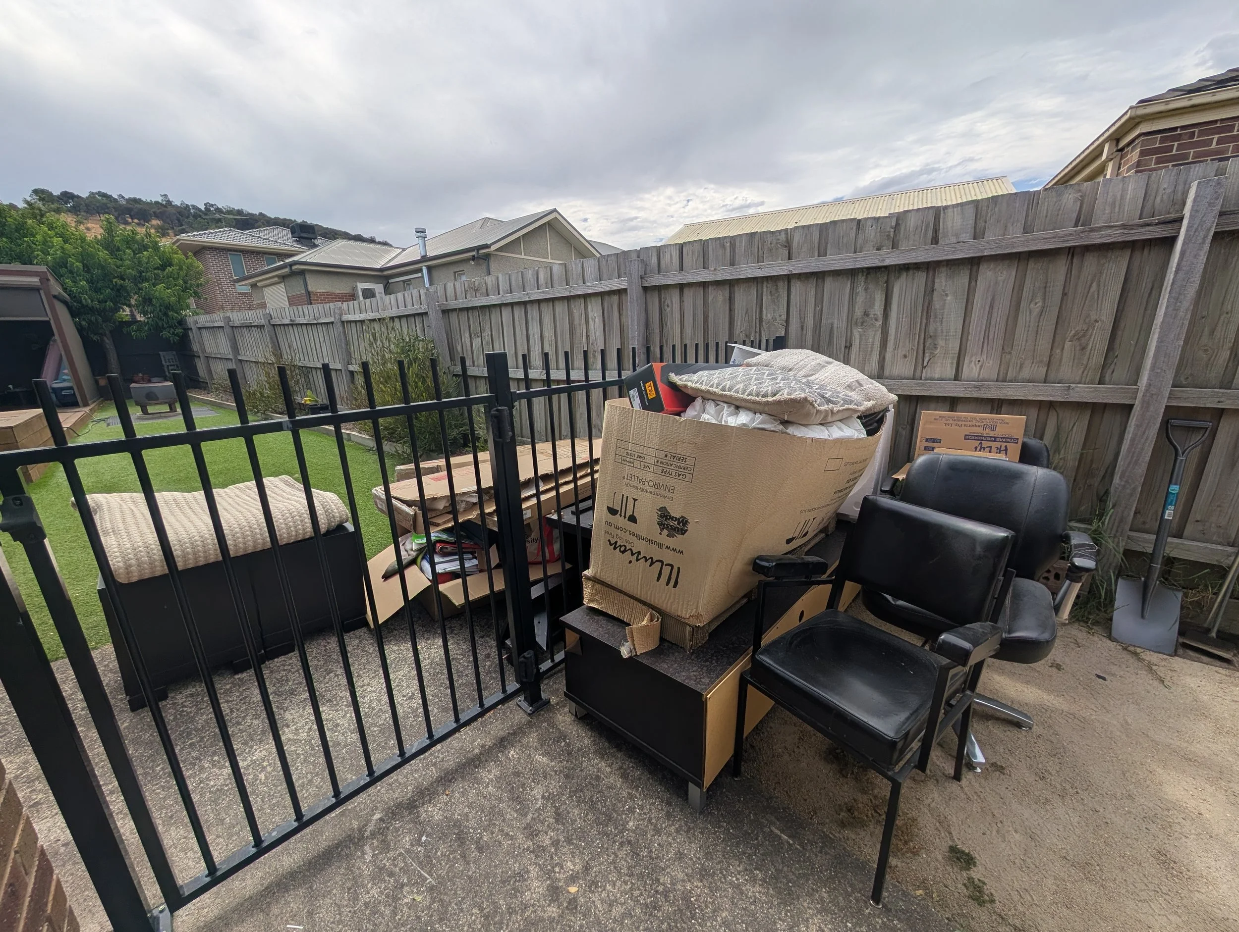 Backyard with a wooden fence, cluttered furniture and boxes, including a cardboard box and a black office chair, on a concrete patio with a grassy lawn and neighboring houses in the background.