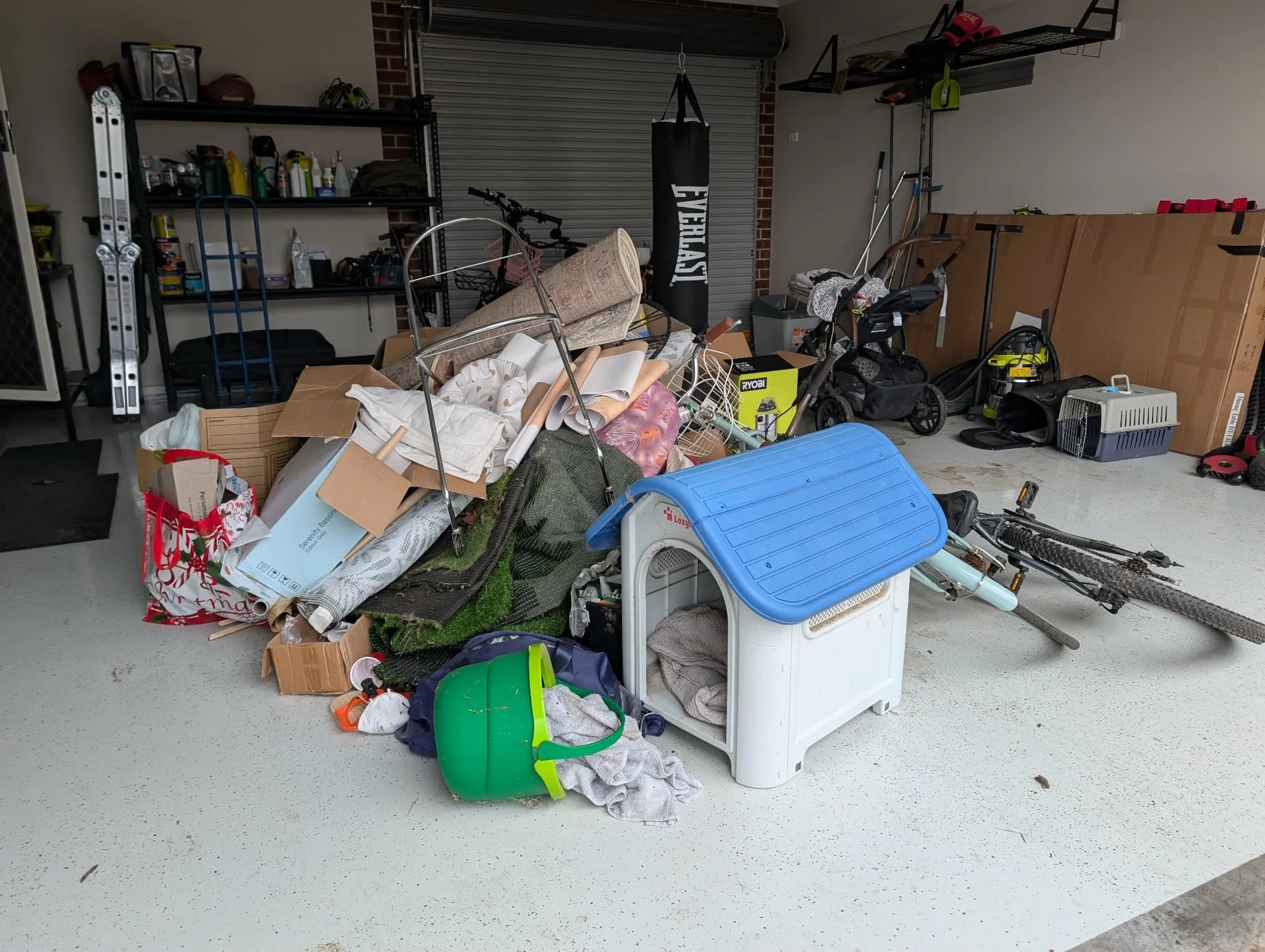 Garage cluttered with various items including boxes, a bicycle, a stroller, a punching bag, garden tools, a pet carrier, and a large cardboard box. Shelves on the wall hold bottles, containers, and miscellaneous objects.