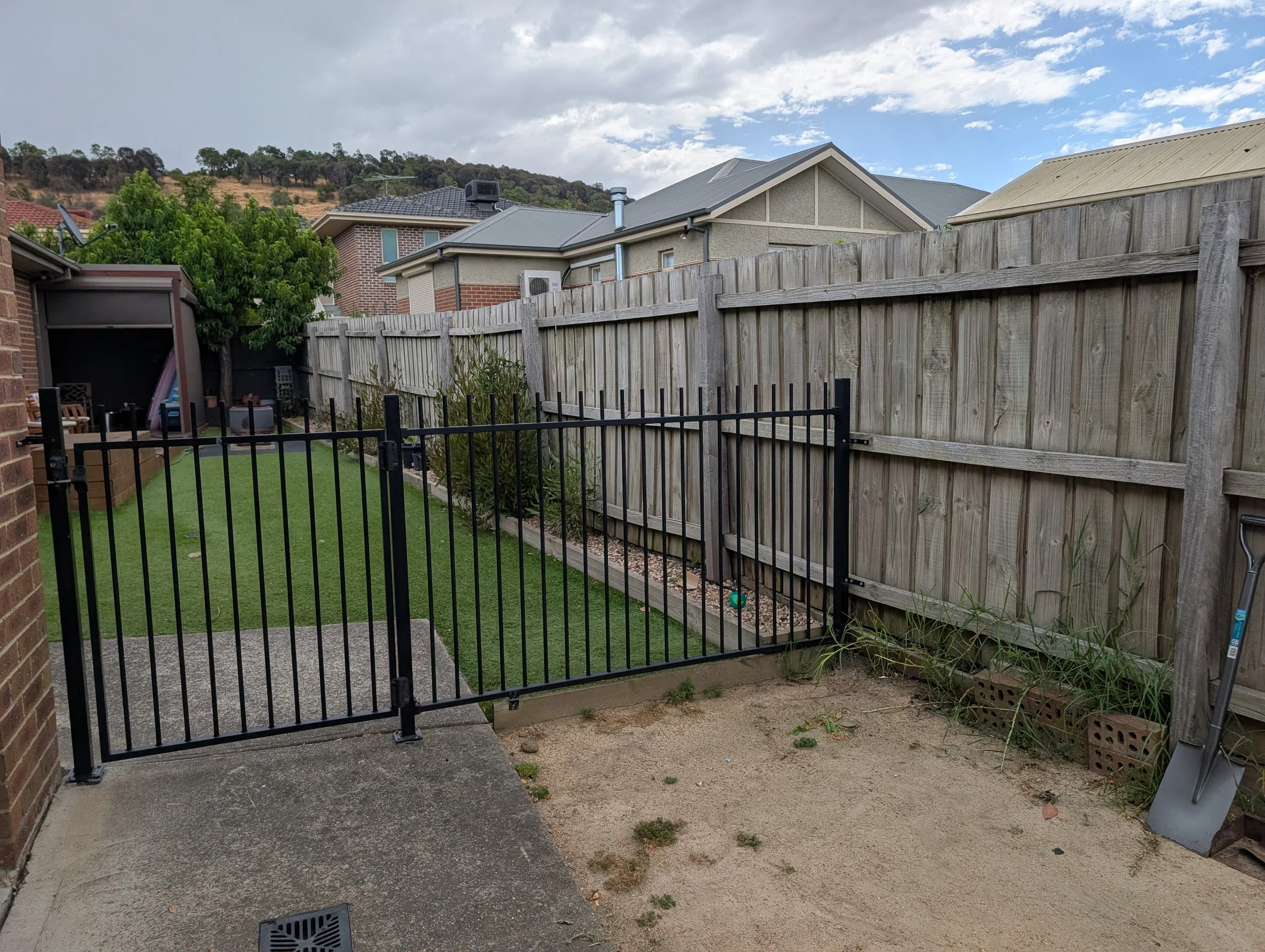 A backyard with a black metal gate, a small artificial grass area, a brick wall, a wooden fence, a garden bed with plants, a metal watering can, and neighboring houses with sloped roofs. The sky is partly cloudy with some blue visible.