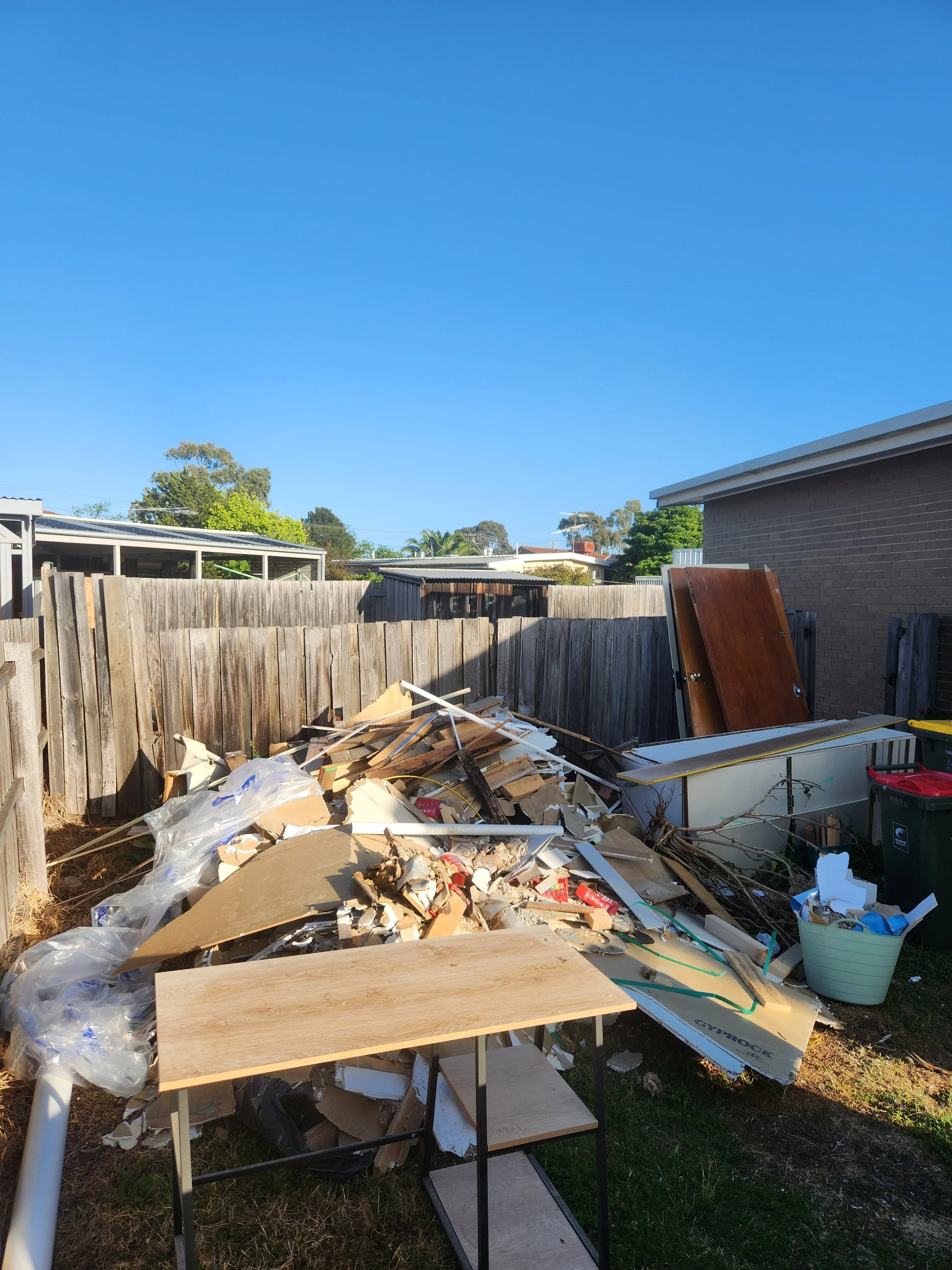 Backyard filled with construction debris, including wood, plastic, and cardboard, with a wooden fence and neighboring houses in the background.