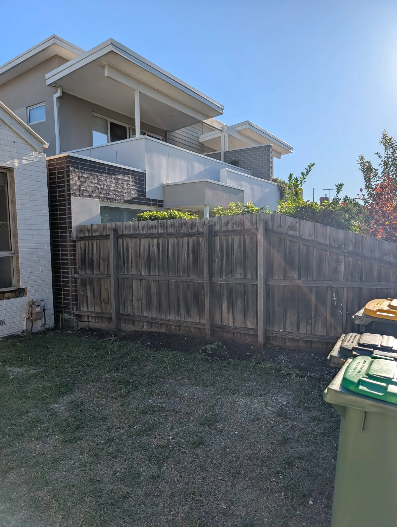 Backyard with a tall wooden fence, grassy patch, some bushes, and a modern multi-story house with balconies under a clear blue sky.