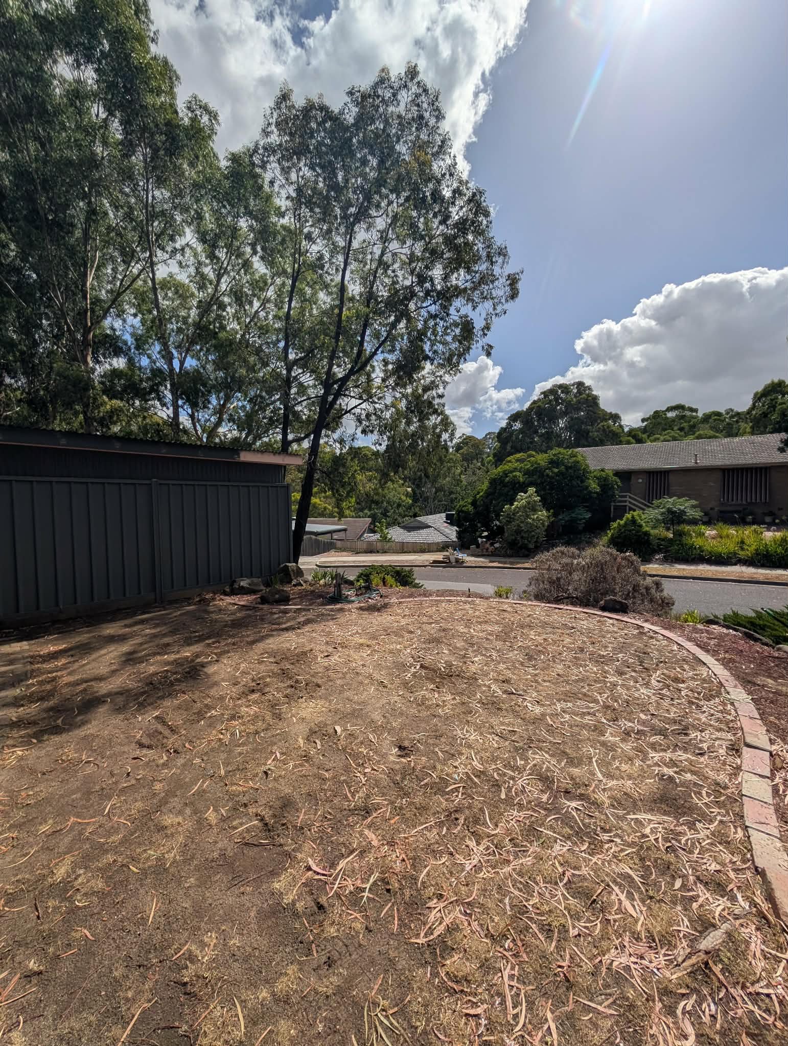 A backyard with a dirt patch and scattered fallen leaves, bordered by a curved brick edge, with a large tree and shrubbery, and neighboring houses in the background under a partly cloudy sky with sunlight.