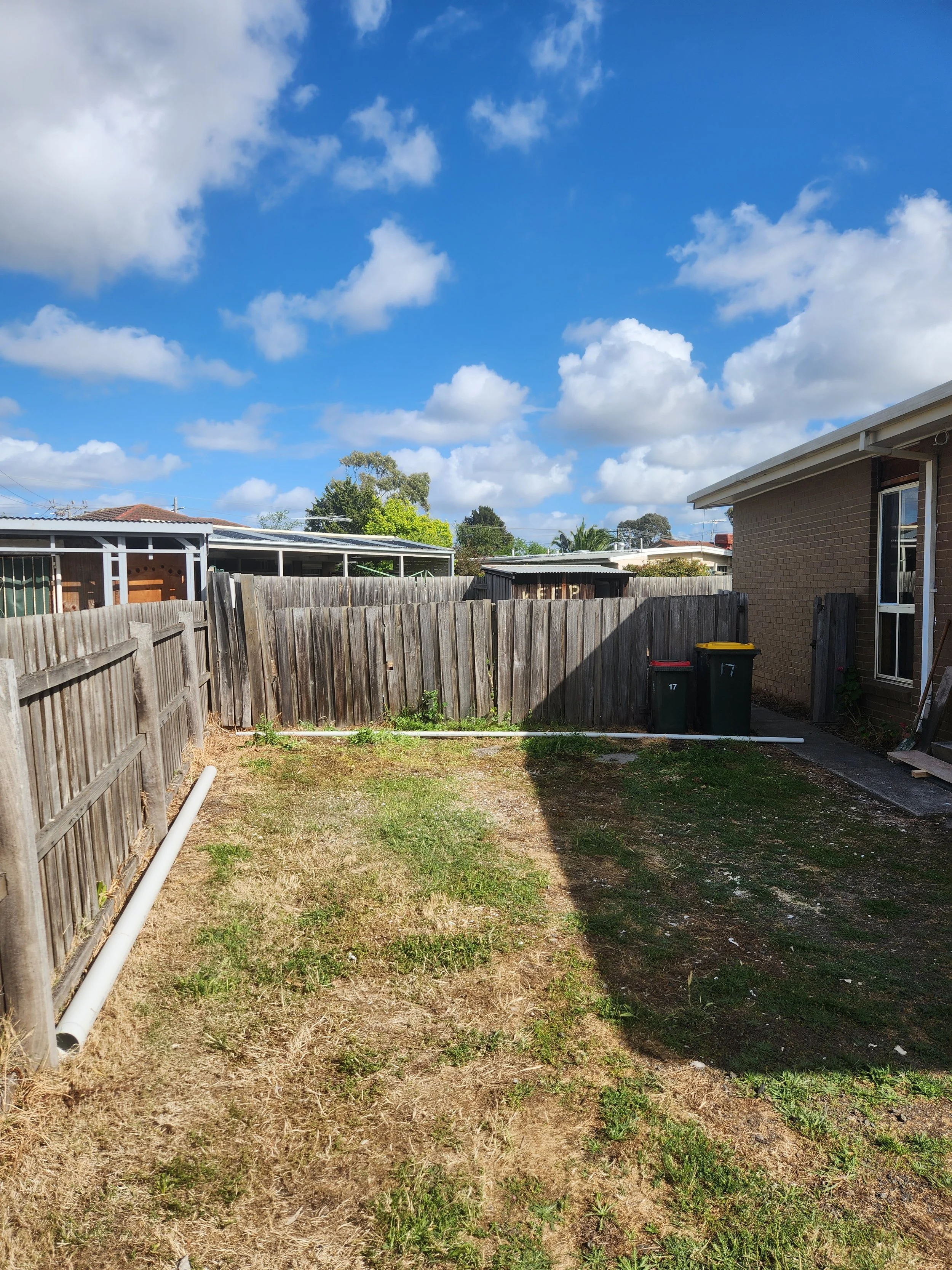 Backyard with a wooden fence, a patch of grass and dirt, two trash bins, a brick house with a window, and a clear blue sky with scattered clouds.