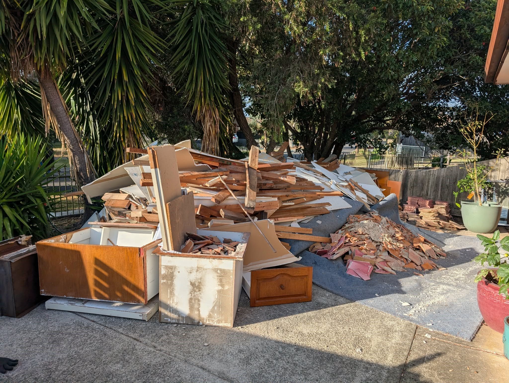Pile of broken furniture and debris outside surrounded by trees and plants.