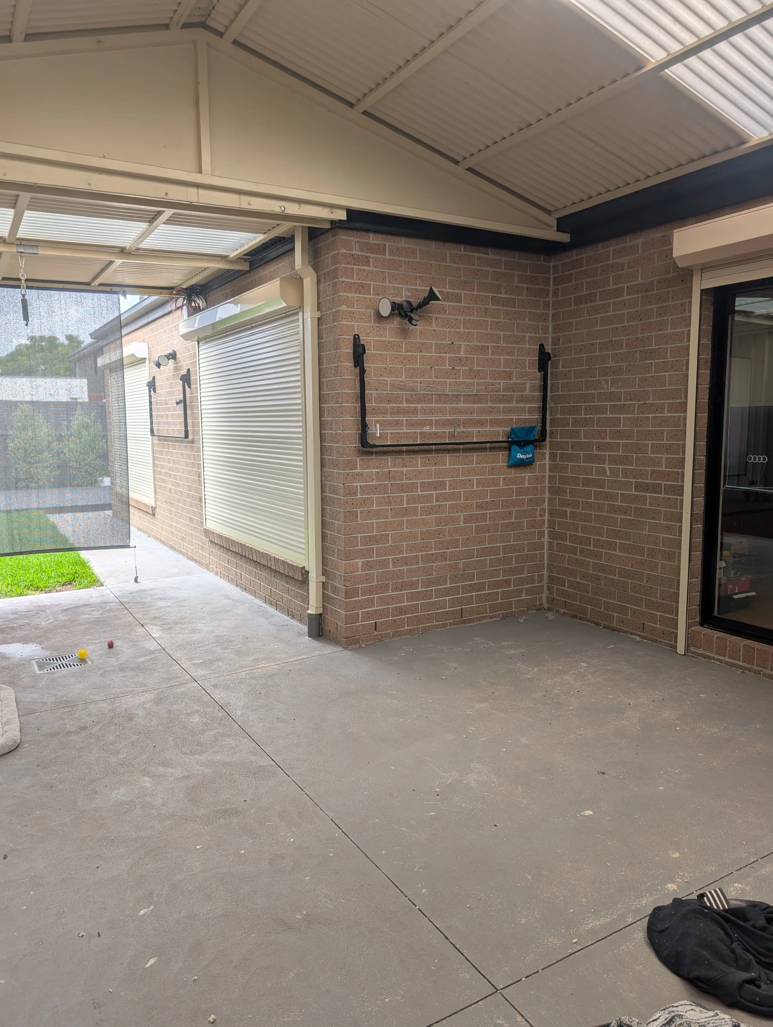 View of an outdoor patio area with a concrete floor, brick wall with a window and black metal pipe, two lawn bowls on the ground, and a black bag in the corner. Part of a screened porch and a hanging wind chime are visible.