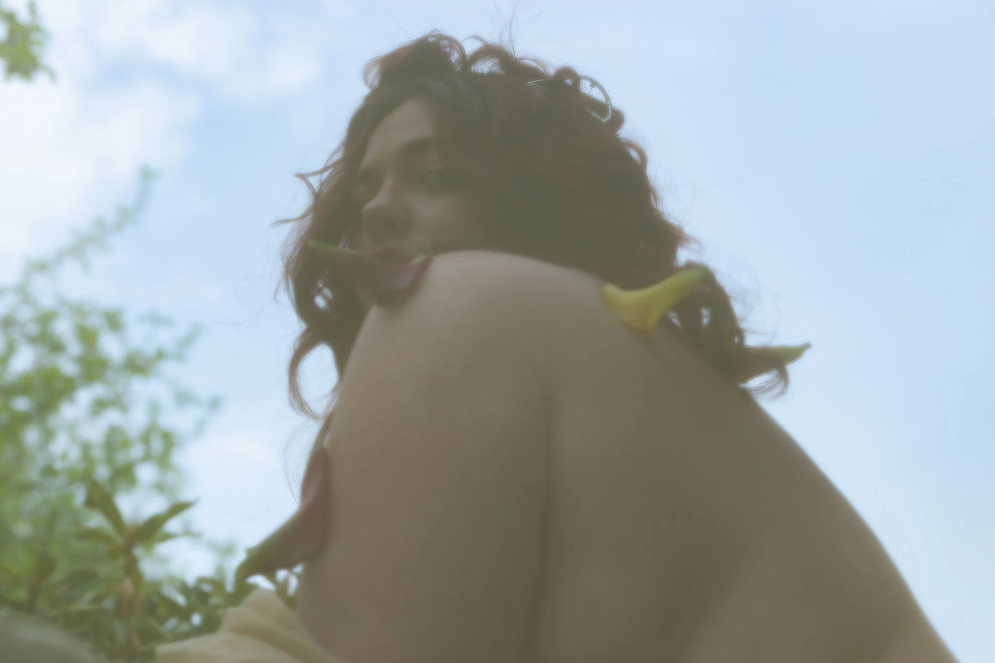 A woman with curly hair is seen from below, looking upward towards a partly cloudy sky with some green foliage in the foreground.