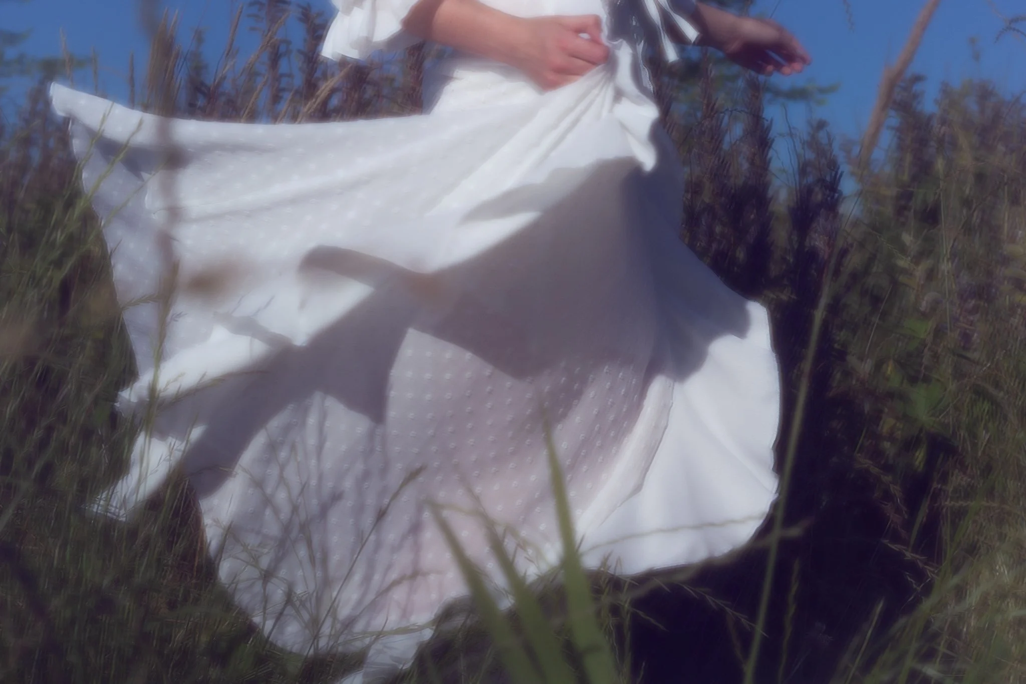 A person dressed in a white standing outdoors in a grassy area with trees in the background.