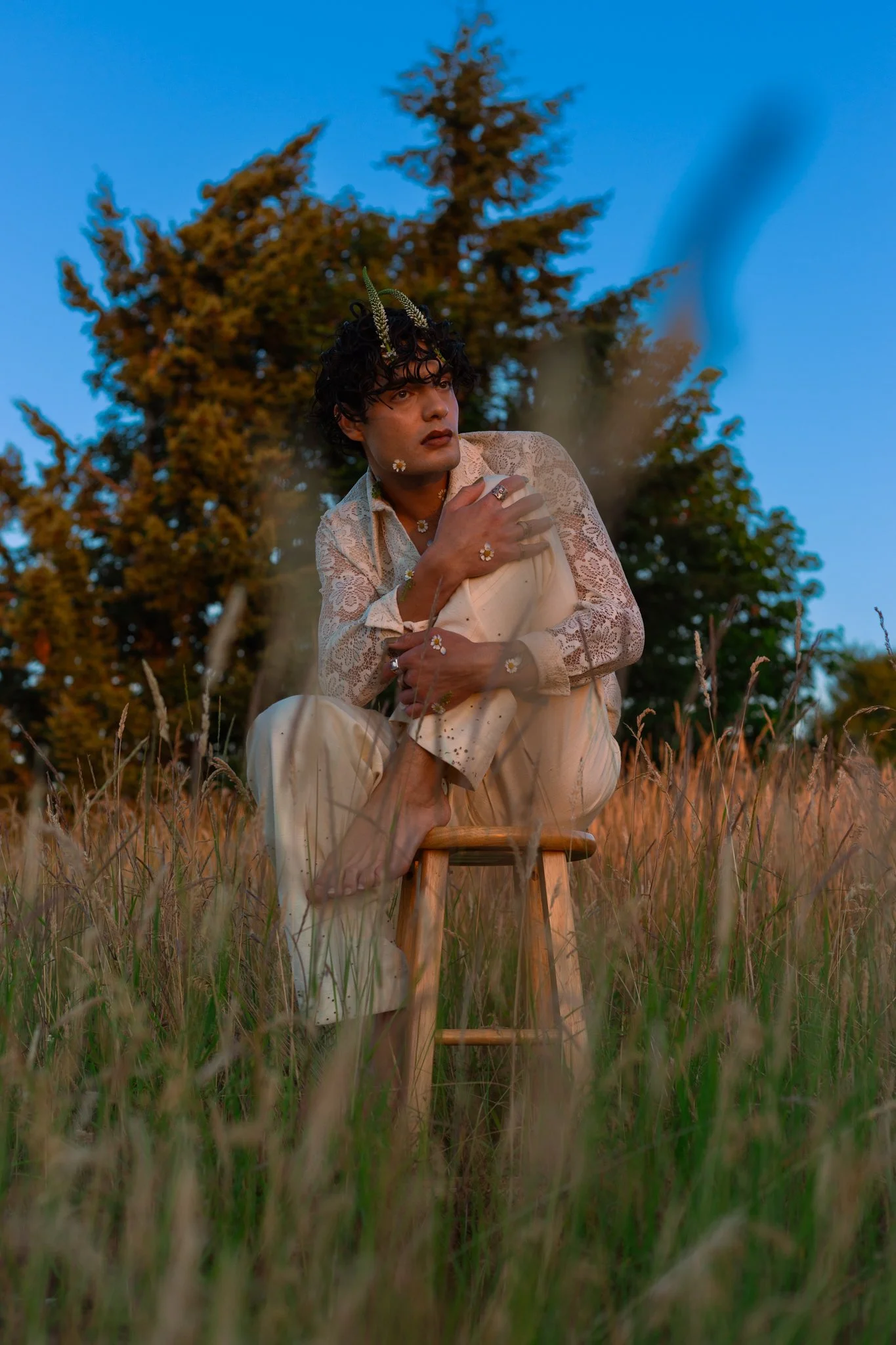 A person with dark curly hair, wearing a white lace shirt and beige pants, is squatting in a field of tall grass during sunset, holding a small stick and looking pensively into the distance. They have flowers on their face, hand, and neck, and are we