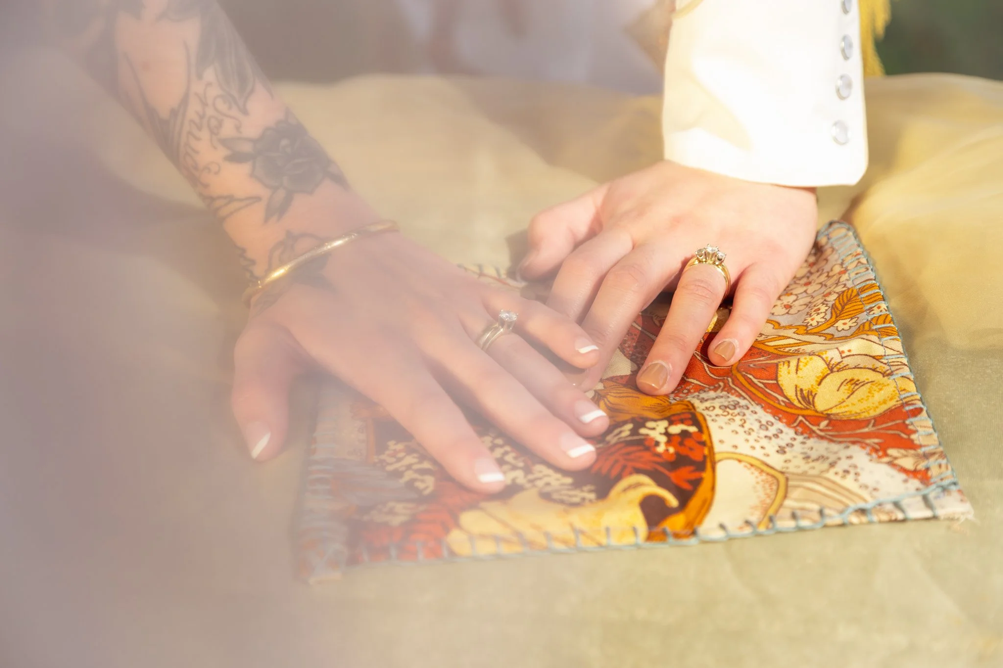 Two hands with rings rest on patterned fabric, one hand has tattoos and both wear rings. Part of white shirt sleeve is visible. The fabric has a colorful, floral and paisley pattern. Blurred background with warm lighting.