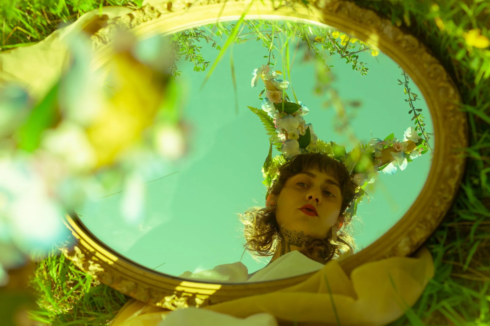 Person looking into a round mirror placed on green grass, wearing a floral headpiece and white clothing, with the sky visible in the reflection.