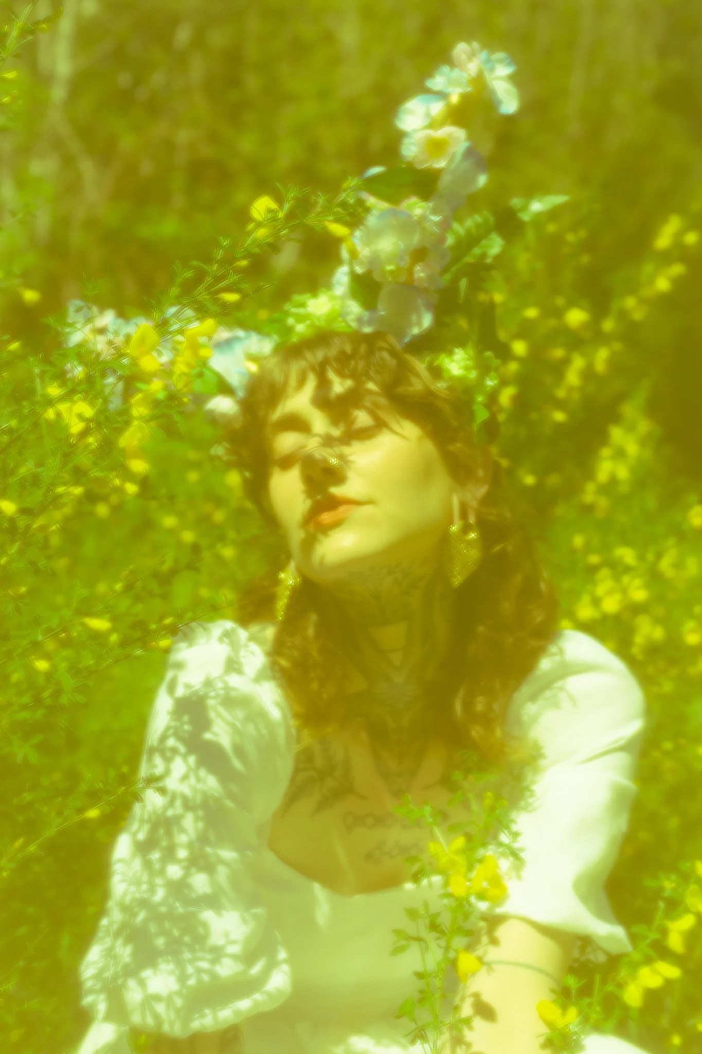	
A person with dark hair and tattoos, wearing earrings and a dark sleeveless top, is standing among green trees and yellow flowers, with floral horns adorning their head