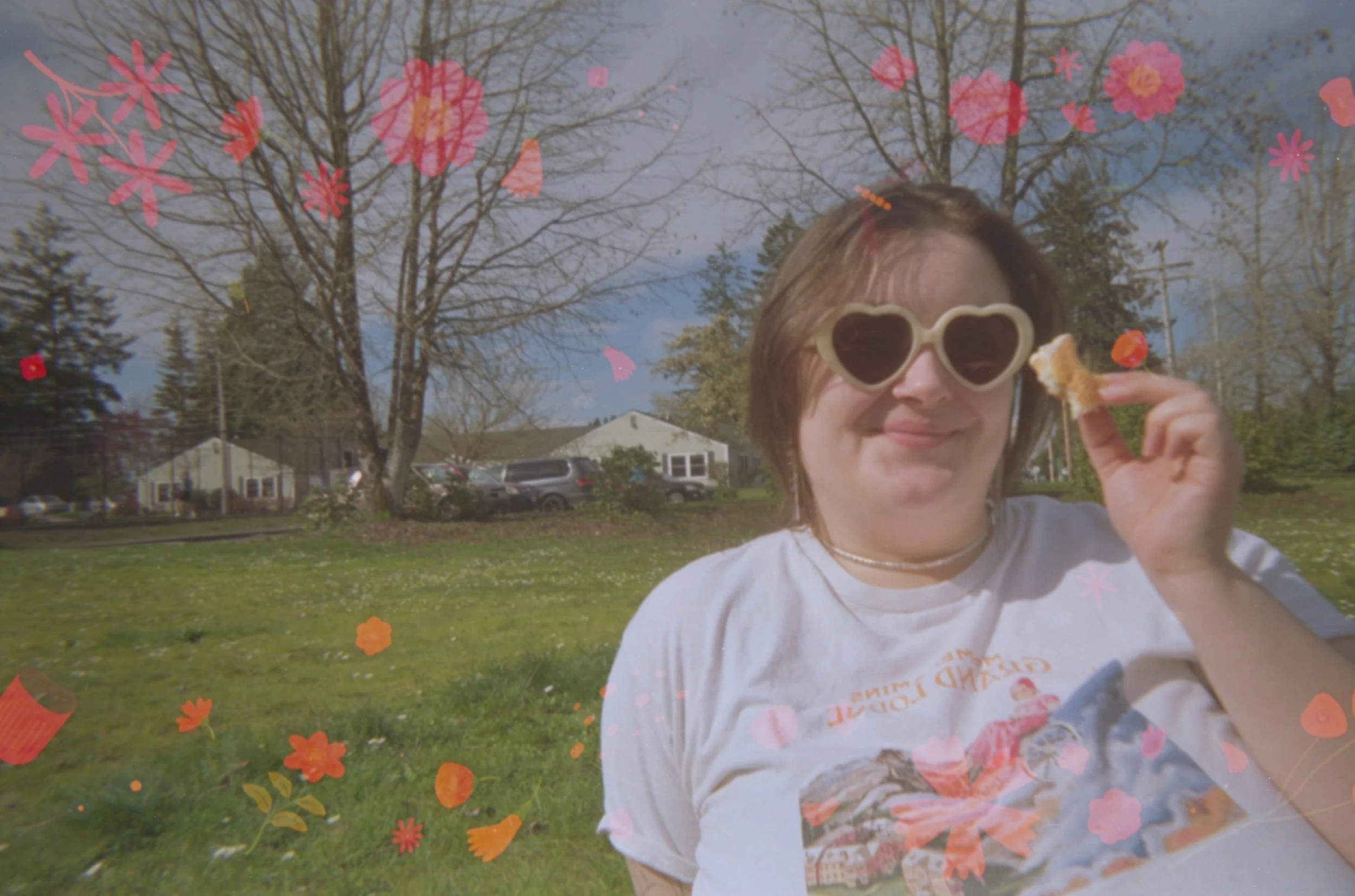 Person with short hair wearing heart-shaped sunglasses and a white t-shirt, holding a piece of food, sitting outdoors with leafless trees and houses in the background. The image has added pink and orange floral overlays.