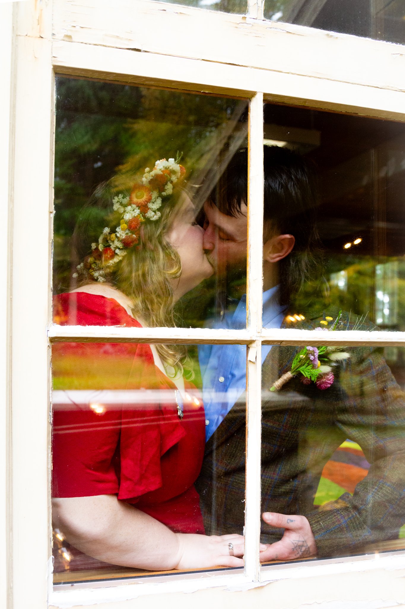 A couple kissing through a window, with the woman wearing a red dress and the man wearing a plaid jacket, holding hands, with flowers in the background.