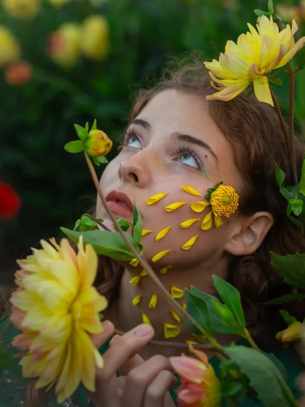 A woman with curly brown hair and blue eyes surrounded by yellow flowers, some of which are placed on her face and head, creating a floral-themed appearance in a garden setting.