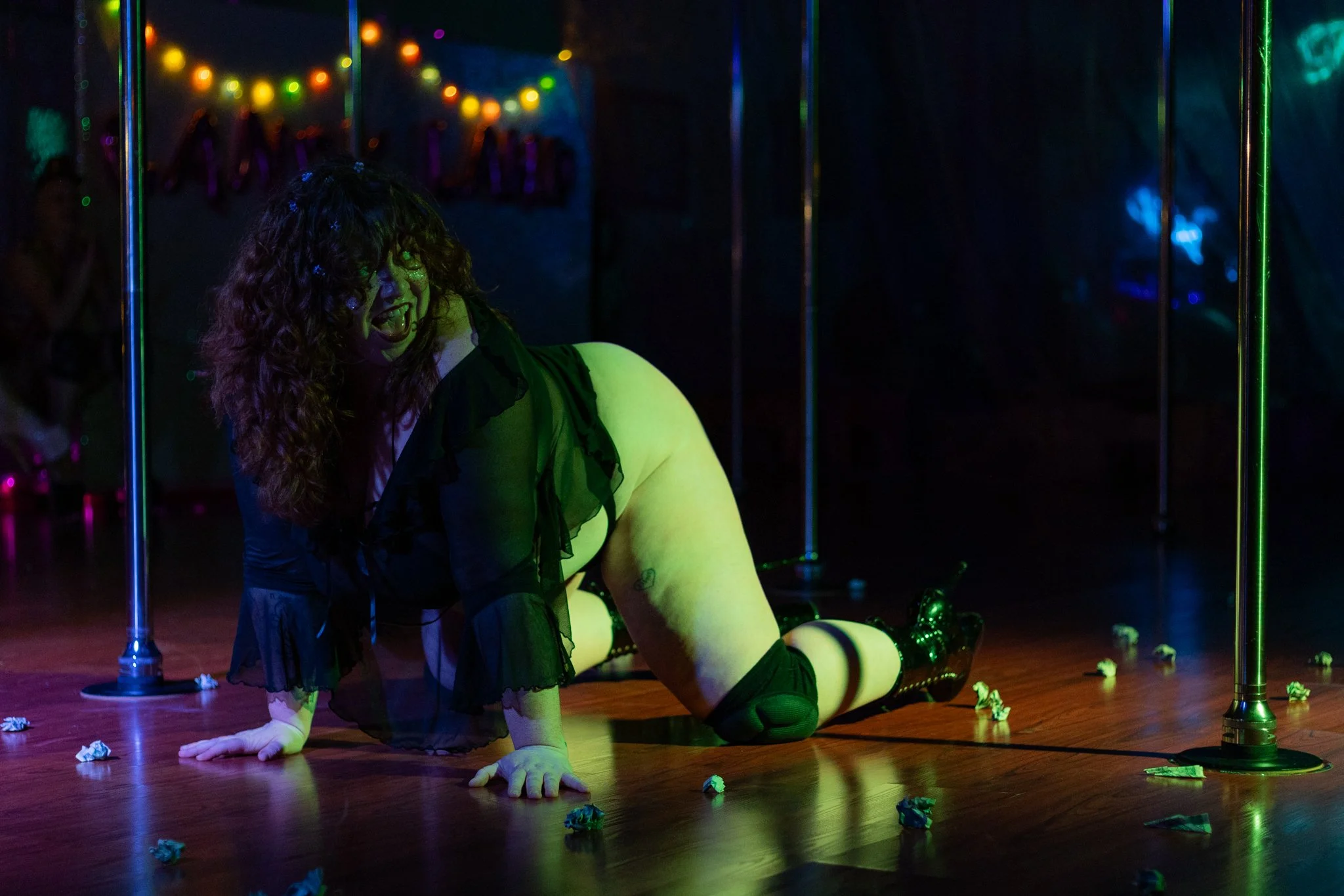 A woman with curly hair performing a pole dance routine on a wooden stage with scattered crumpled paper. She is wearing black lingerie, thigh-high stockings, and gloves, with a colorful light illuminating her and the background. The scene appears to be in a nightclub or performance venue.