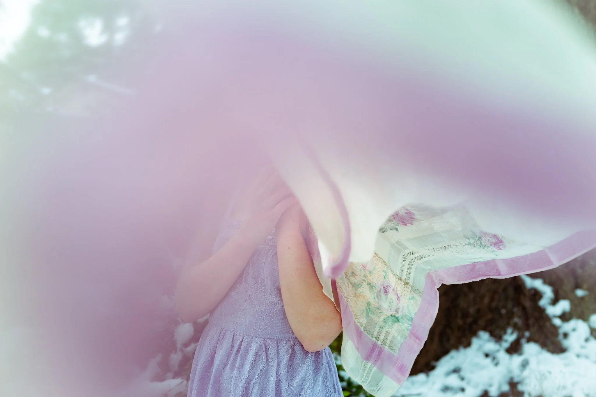 A person wearing pastel-colored dresses. The person is partially covered by a pink fabric, and the background includes some greenery and dirt.