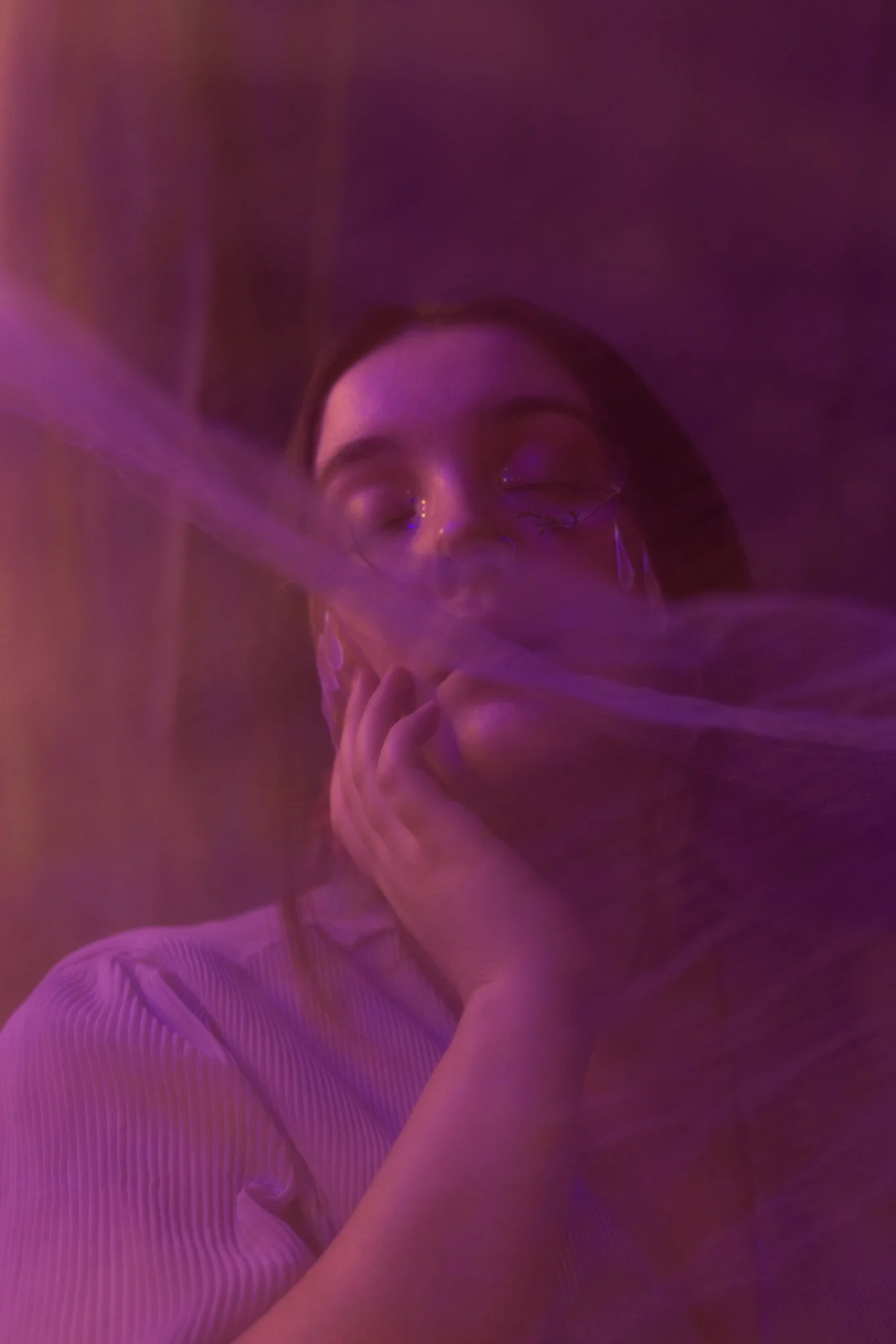 A person with glasses and earrings, wearing a light-colored, textured shirt, appears to be peacefully resting with eyes closed, surrounded by a dreamy, purple-hued mist or smoke.