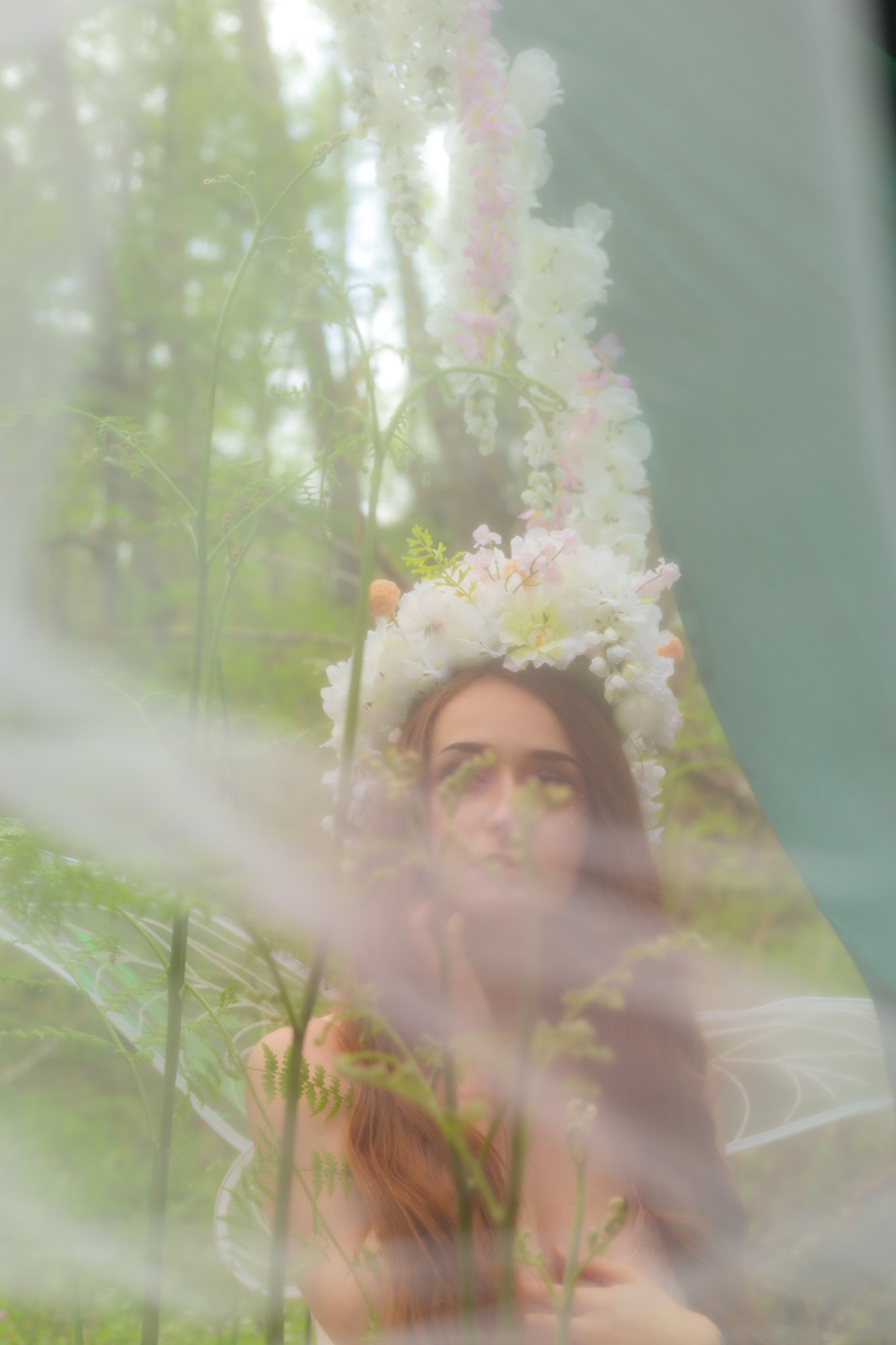 A woman with long red hair wearing a floral crown with white and pink flowers, seen through a fabric with green foliage around.