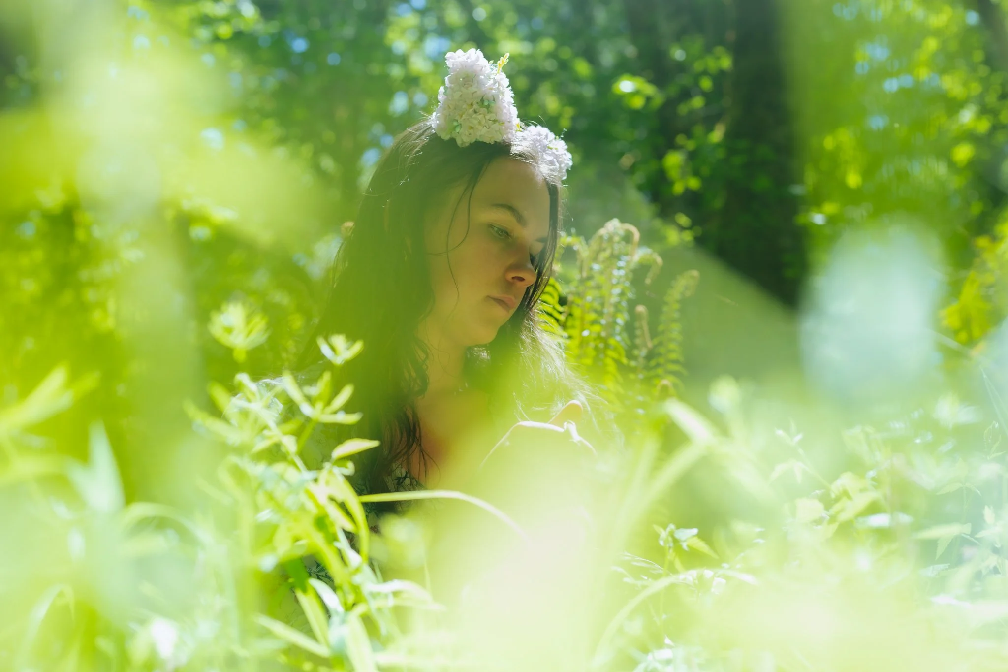 A young woman with long dark hair, wearing a floral crown, is looking down in a lush green forest with sunlight filtering through the trees.
