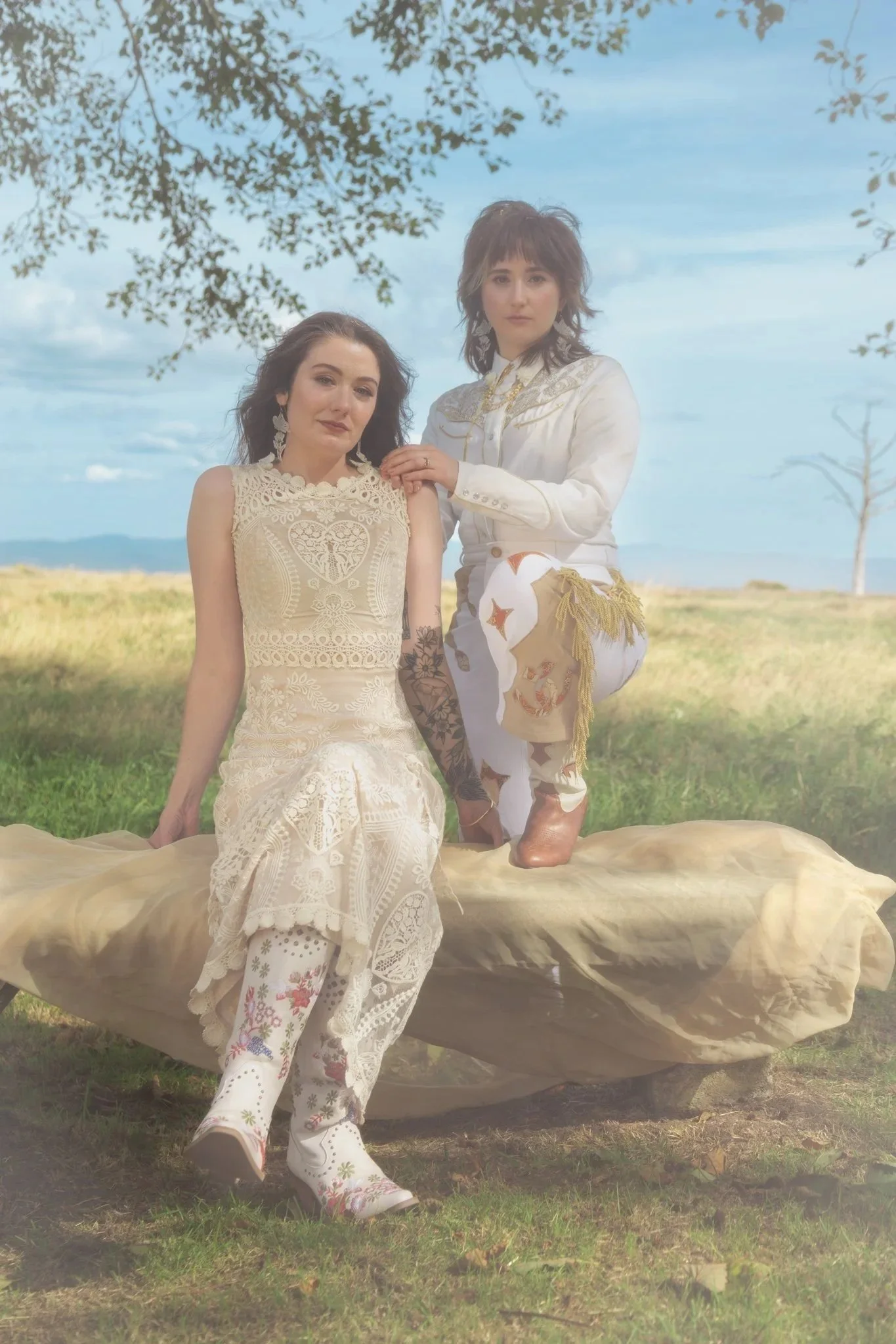 Two women outdoors, one sitting on a large, flat stone and the other standing on it with one foot, both wearing light-colored, ornate clothing, with trees and blue sky in the background.