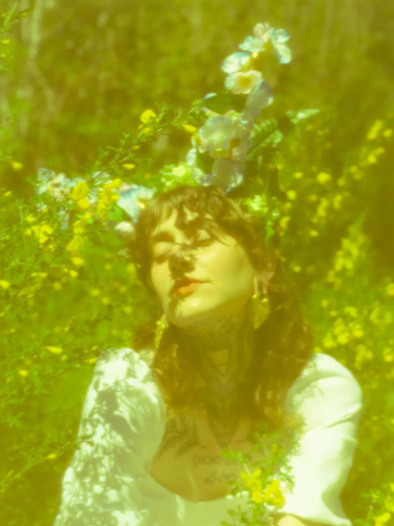 A person with dark hair and tattoos, wearing earrings and a dark sleeveless top, is standing among green trees and yellow flowers, with floral horns adorning their head