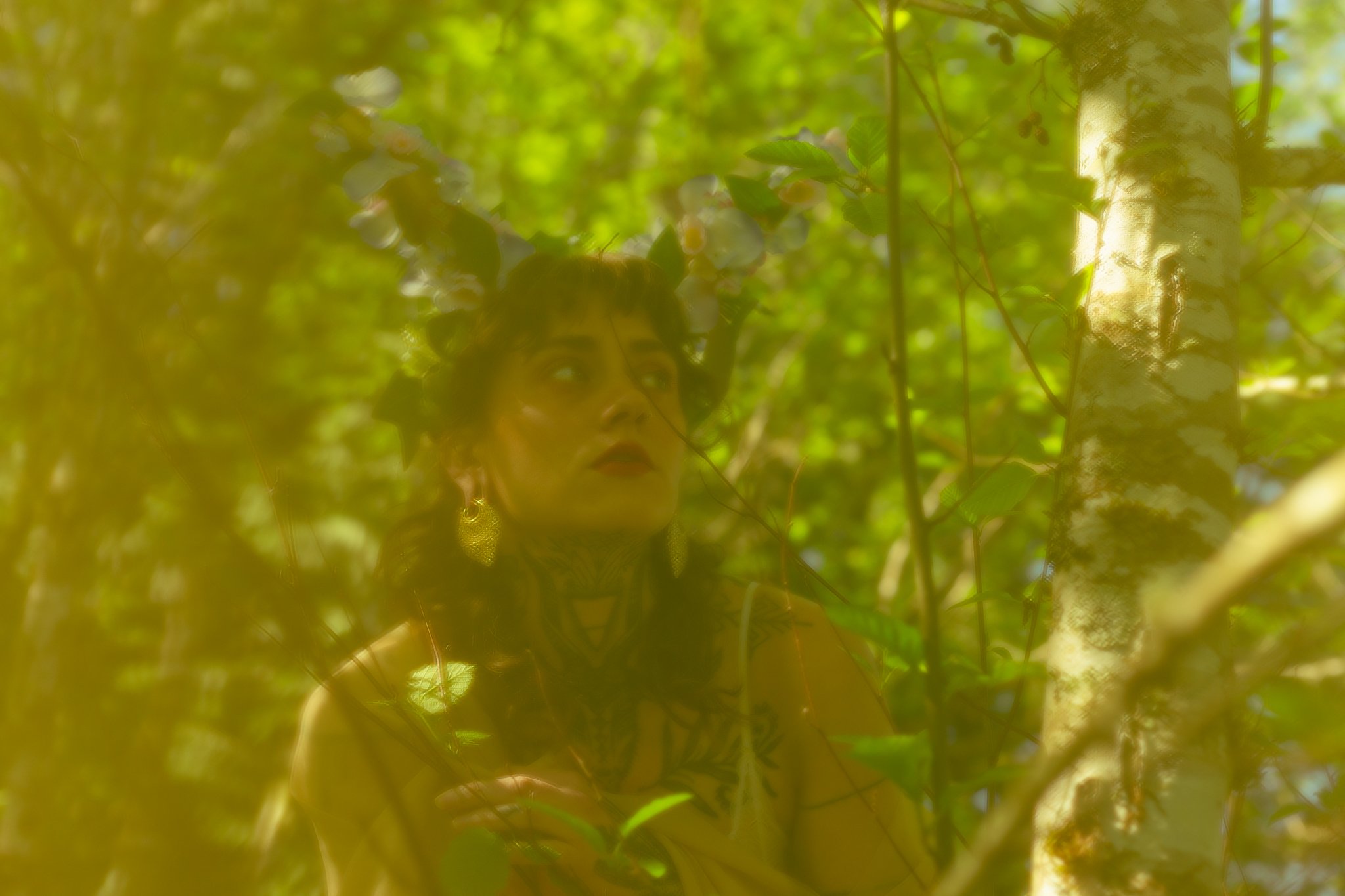 A reflection of a woman wearing jewelry and a hat in a mirror surrounded by green leaves, with a tree trunk partially visible.