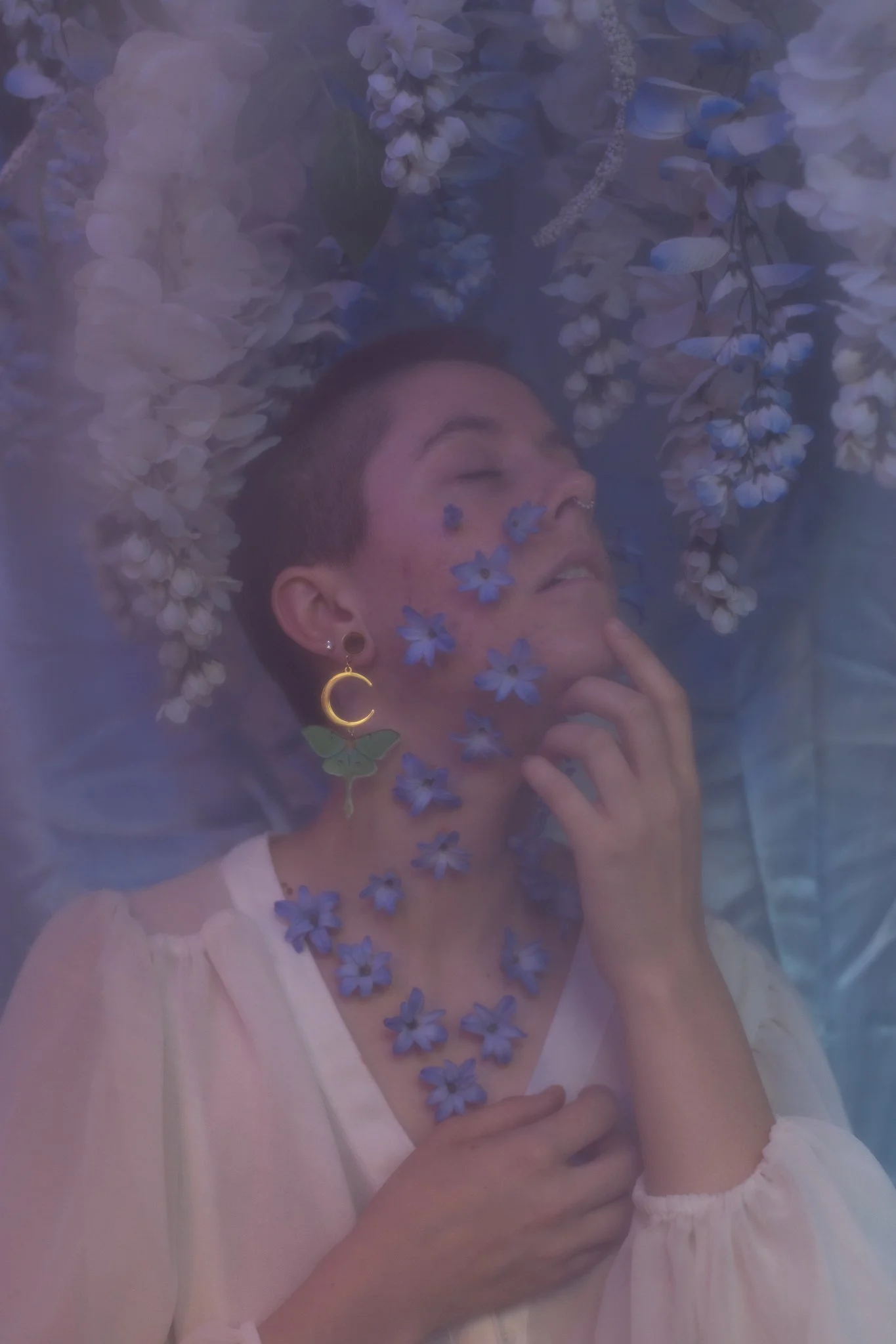 A person with short dark hair surrounded by purple flowers and petals, with eyes closed and a serene expression, wearing a white top and a gold moon earring.