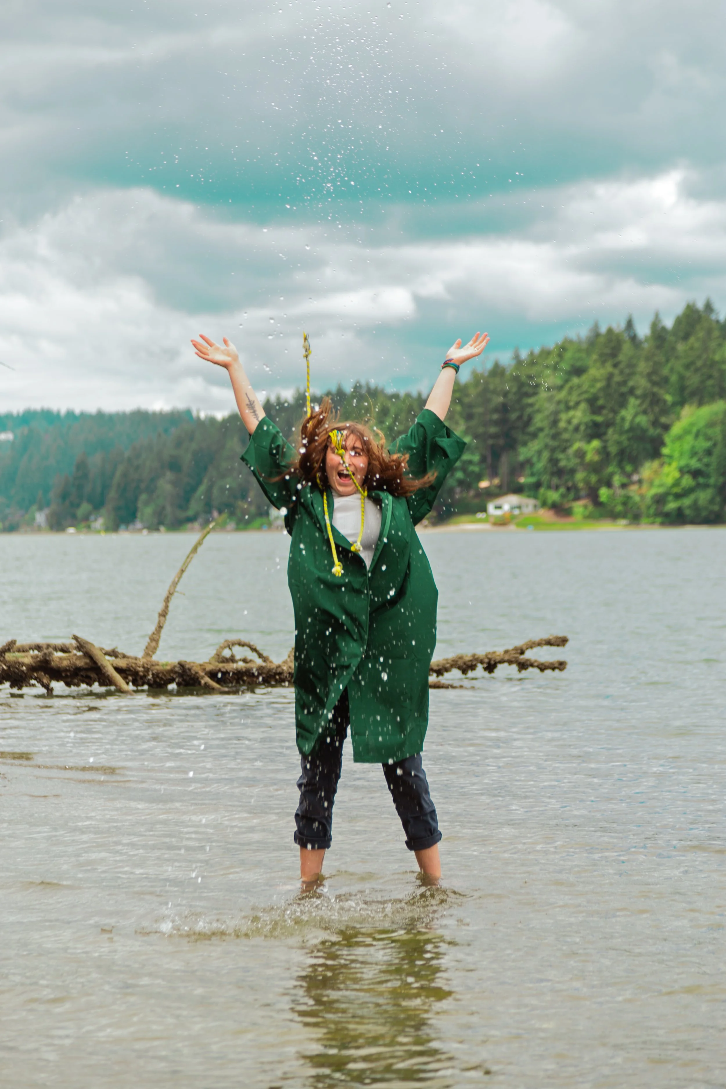 A person standing in shallow water near a tree and riverbank, wearing a green graduation gown and celebrating with arms raised on a cloudy day.